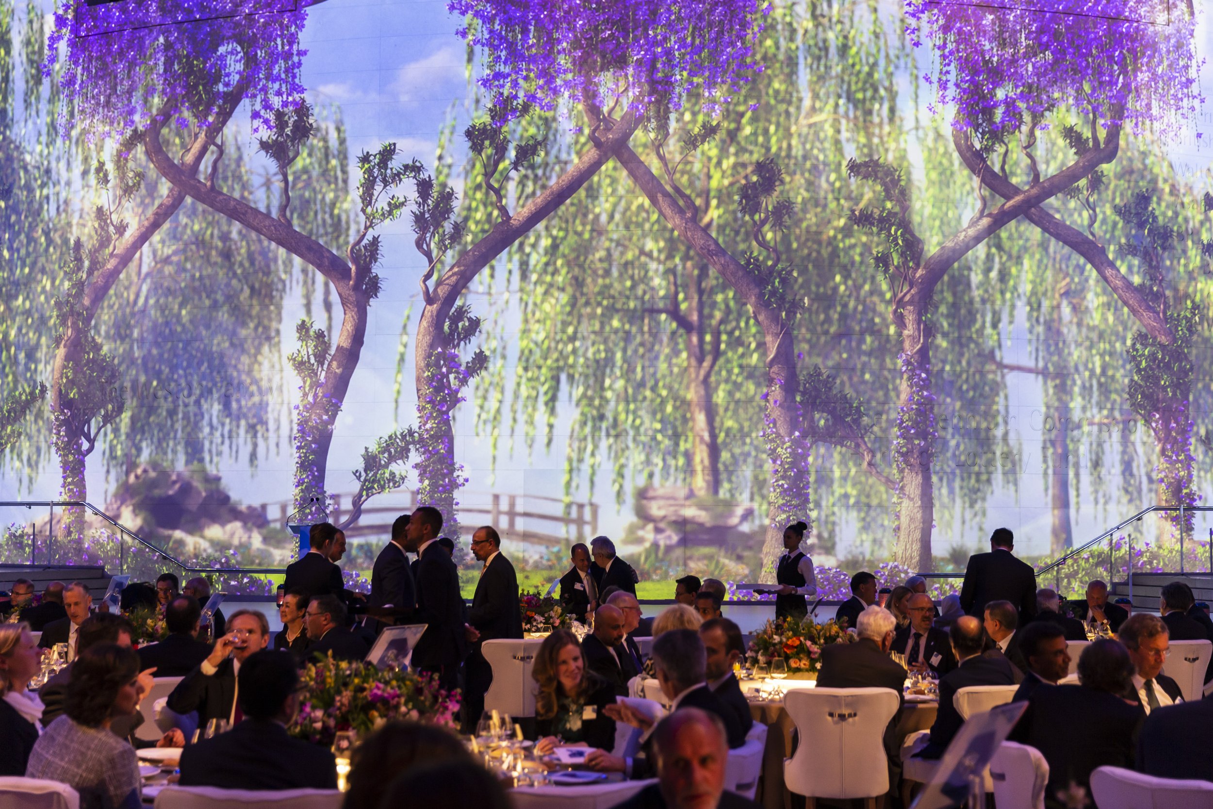 A large banquet hall with many guests seated at decorated tables, some standing and conversing, and a big digital screen displaying a scenic outdoor landscape with trees, flowers, and a blue sky in the background.