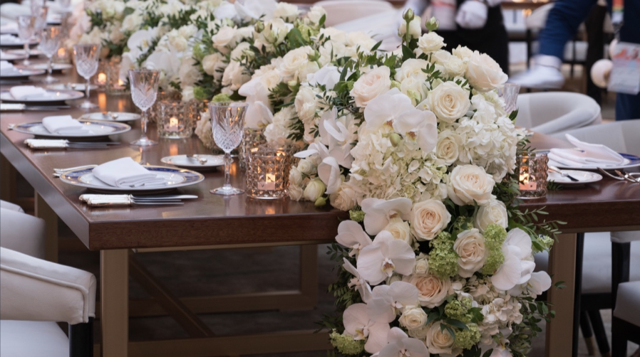 A long wooden dining table decorated with an elaborate arrangement of white roses, orchids, and hydrangeas, with crystal glasses, candles, and white table settings, likely for a wedding or special event.