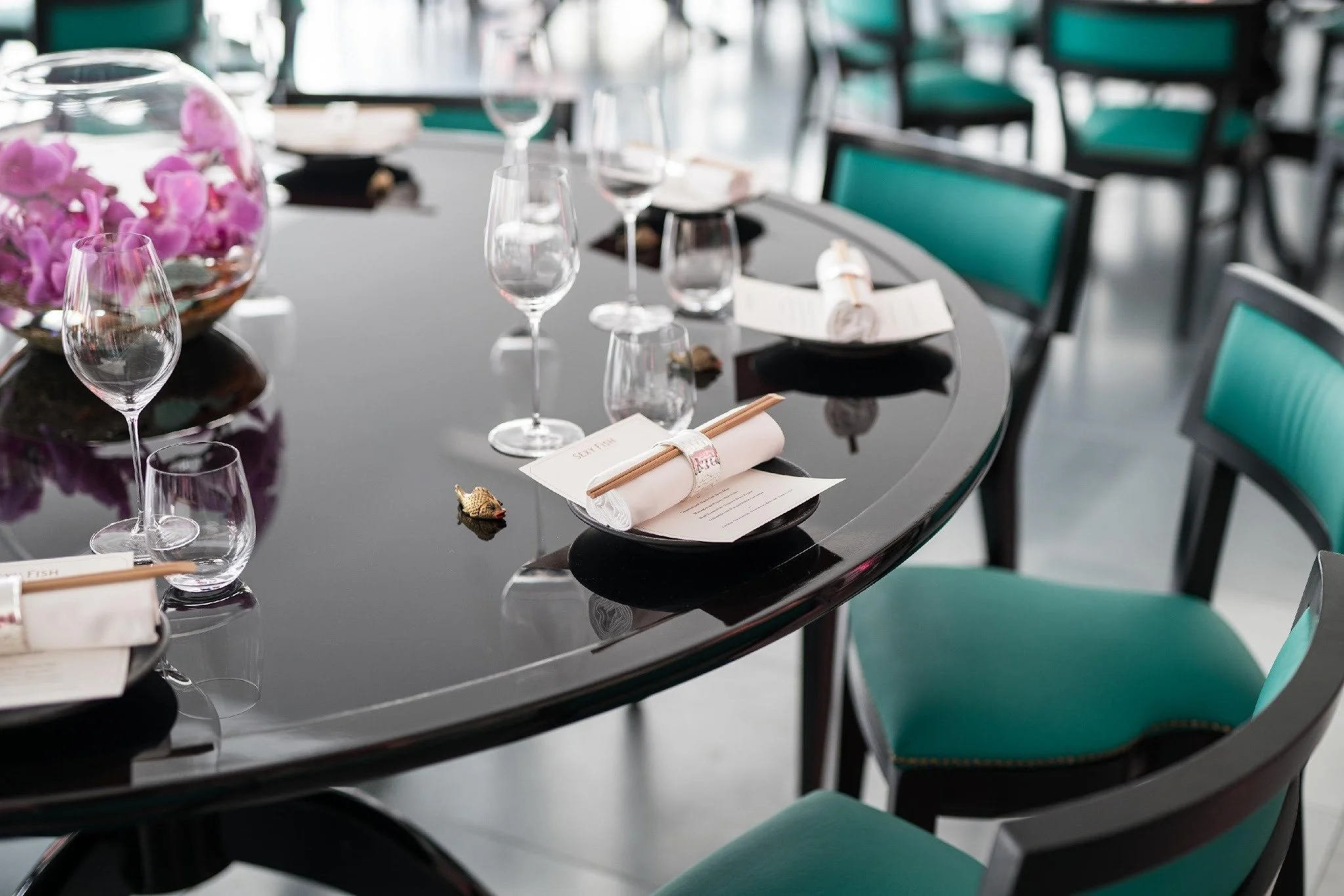 Elegant dining table set with wine glasses, rolled napkins with chopsticks, and a large glass bowl filled with pink flowers in a modern restaurant or event space.