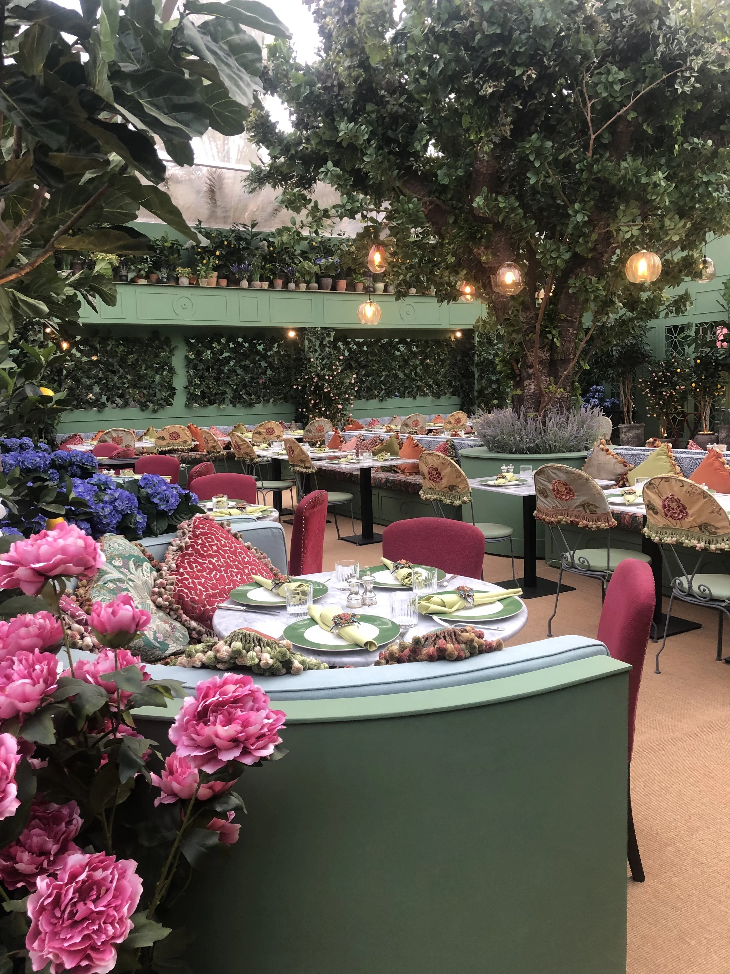Elegant indoor garden restaurant with pink and red floral pillows, colorful table settings, and lush greenery including large trees, plants, and flowers, illuminated by hanging lantern lights.
