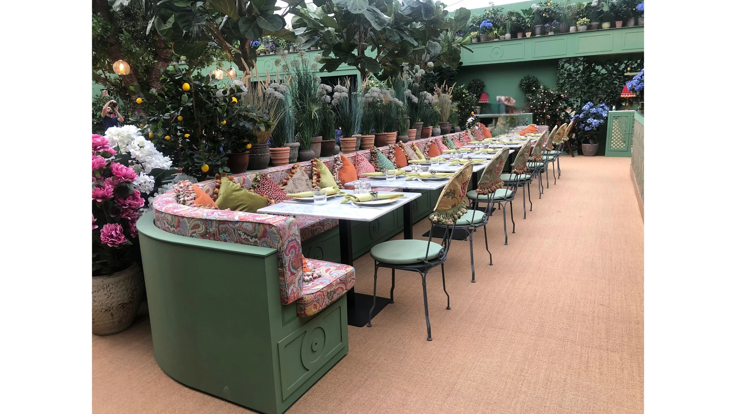 A long dining table set for a meal in a lush, vibrant indoor garden with potted plants and flowers, cushioned chairs, and decorative pillows, with a person taking a photo in the background.
