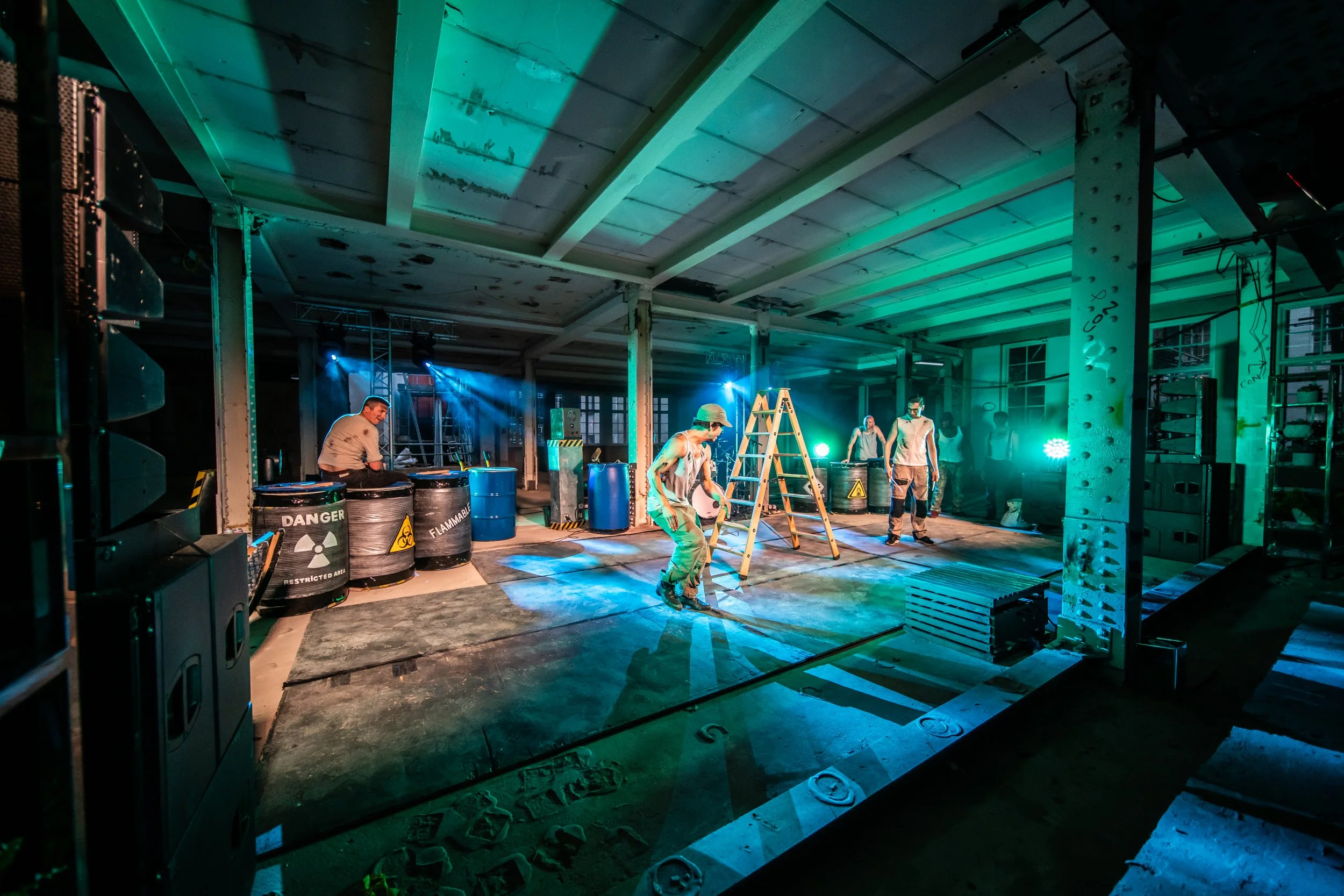 A group of people dressed in protective clothing and helmets working inside an industrial space with blue and green lighting, surrounded by barrels and construction equipment.