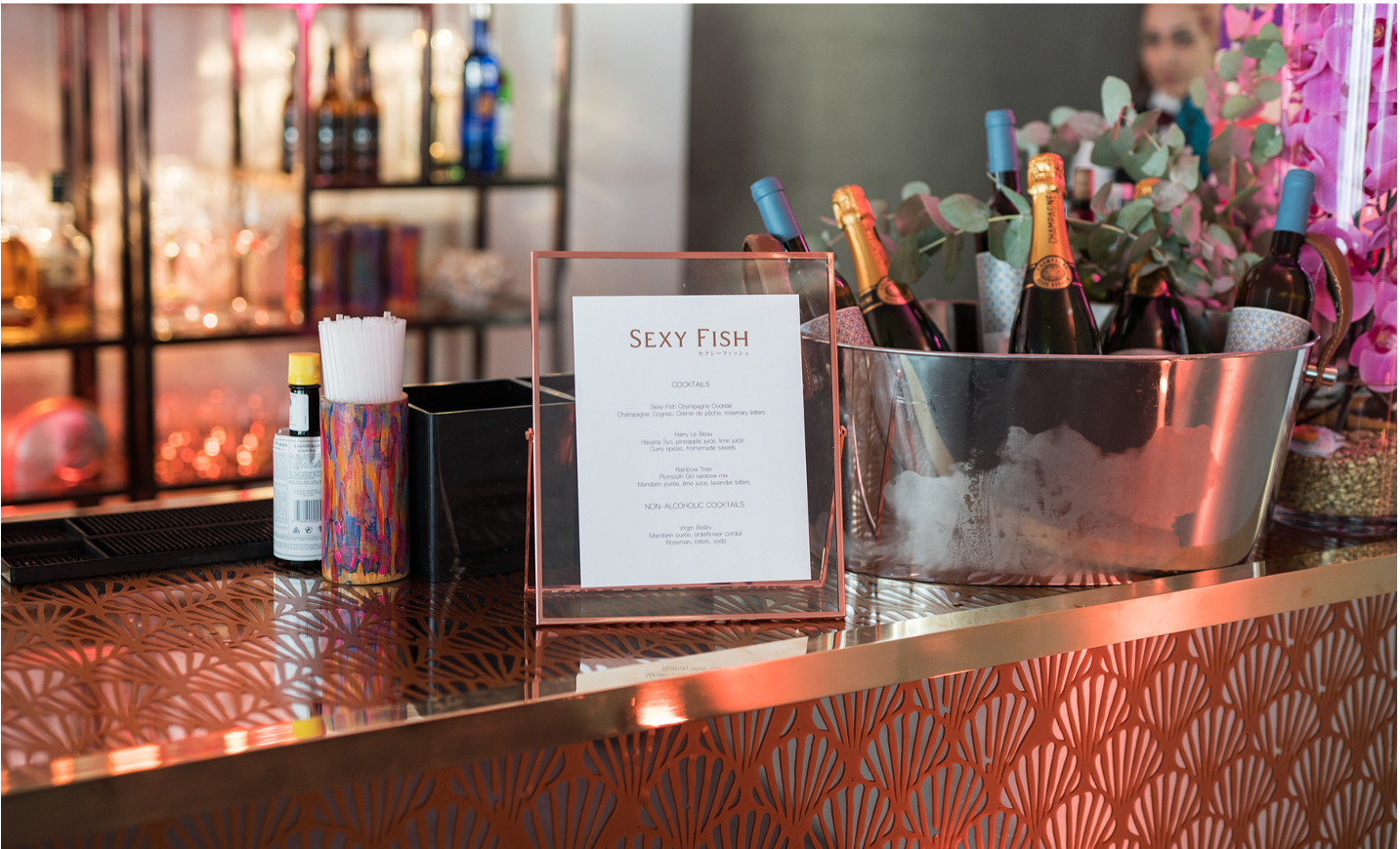 A bar counter with a menu labeled 'Sexy Fish' featuring cocktail options, bottles of champagne and wine, a colorful cup holding straws, and a large metal ice bucket with bottles of champagne and pink flowers.