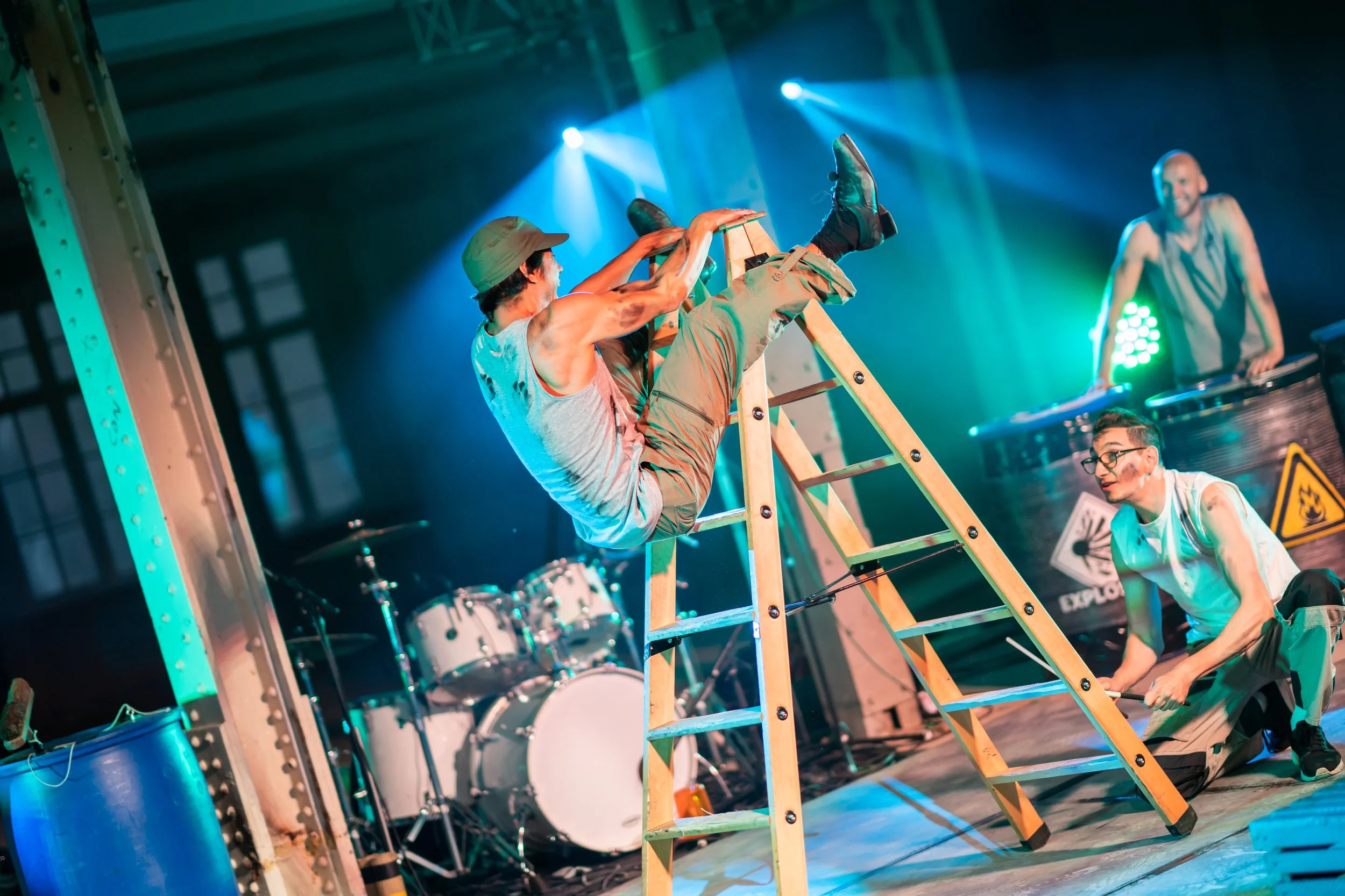 People performing a stunt on stage with a ladder, set up as part of a show, with drum set and stage lighting in the background.