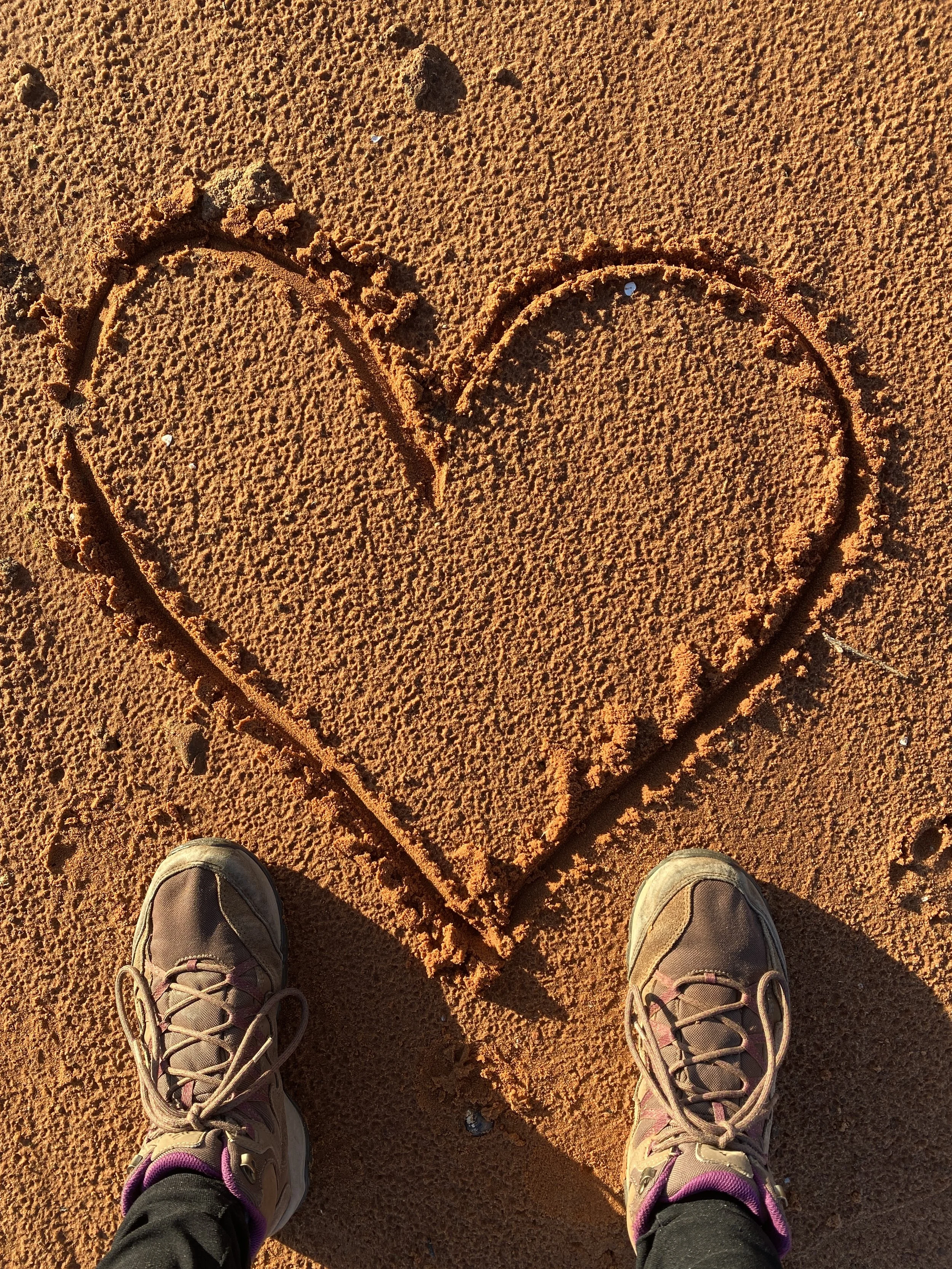 Shoes standing on sand with a heart shape drawn in the sand.