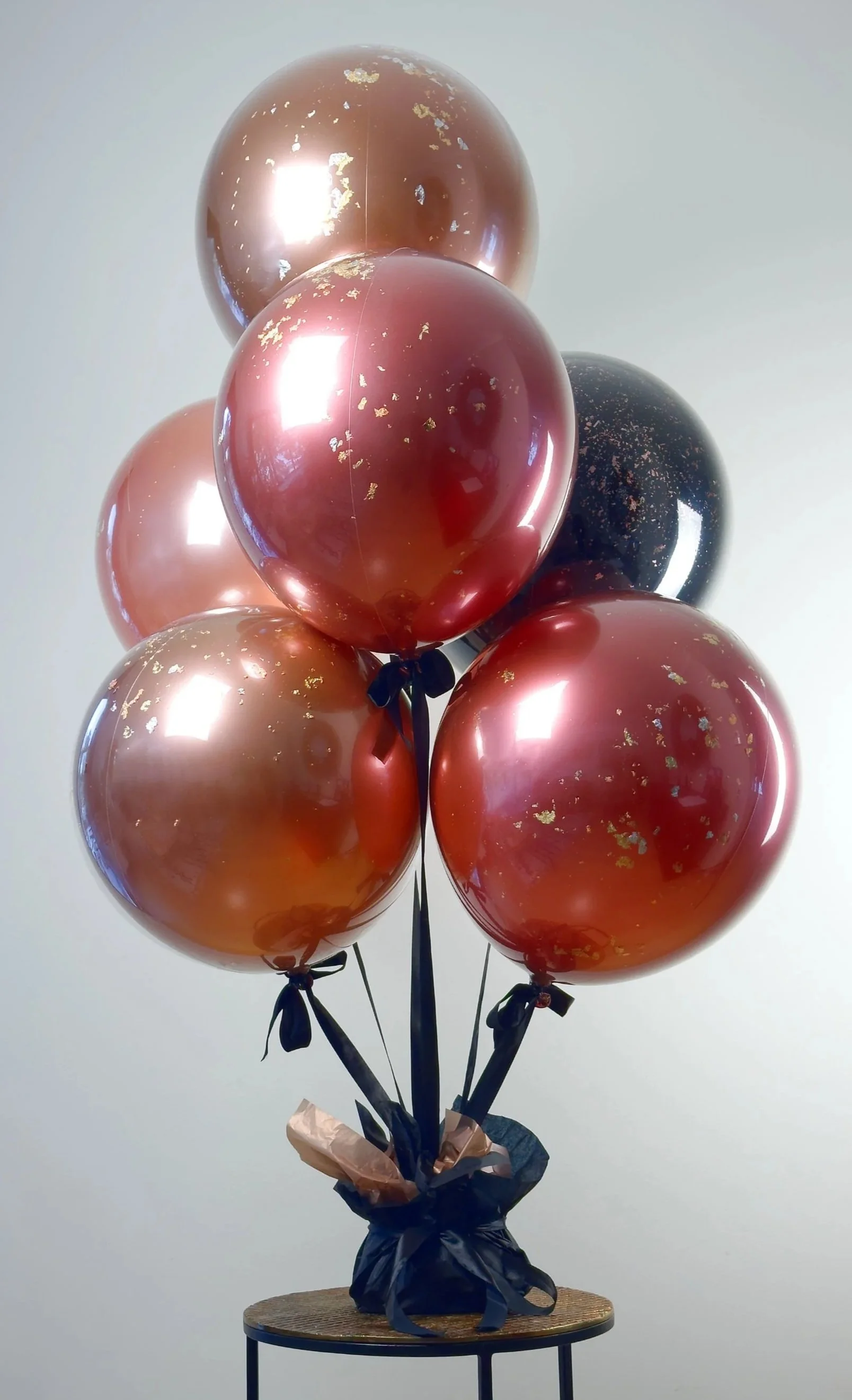 Six metallic balloons in shades of bronze, copper, and black, with some gold flecks, tied together with black ribbons, sitting on a round wooden table against a plain background.