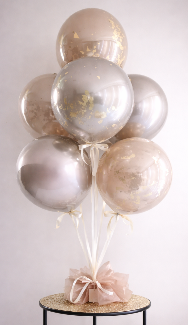 A bouquet of ivory and light pink balloons with gold flecks, tied with white ribbons, arranged on a round table with a gold glitter top and a pink tissue paper base.