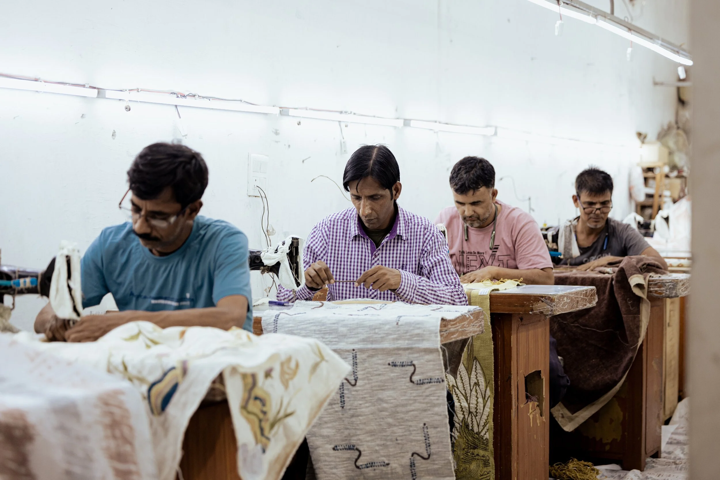 Four men working on sewing or textile projects at a workshop or factory, sitting at tables with fabrics and sewing machines.