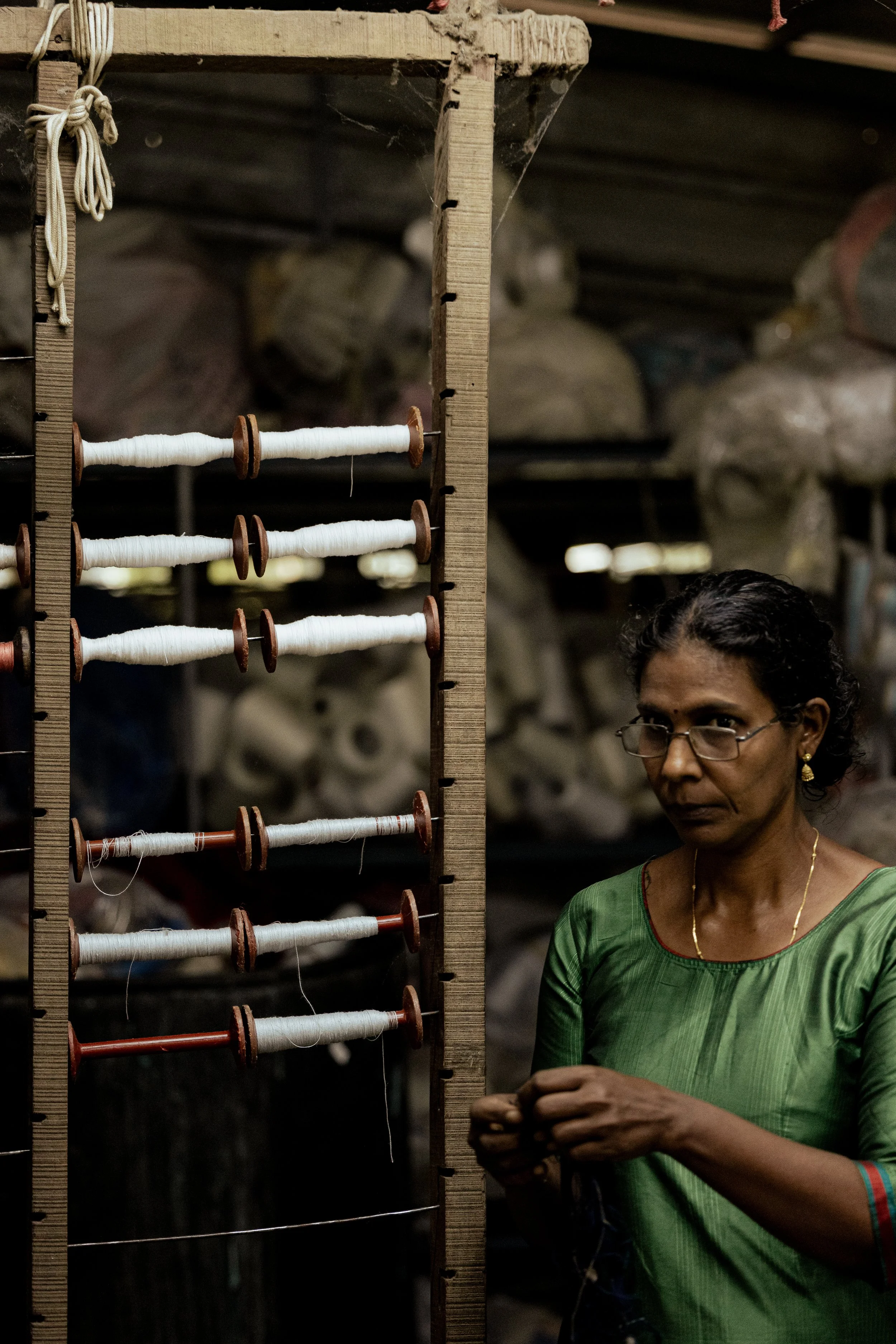 A woman in a green saree is working with threads on a wooden frame in a textile workshop.