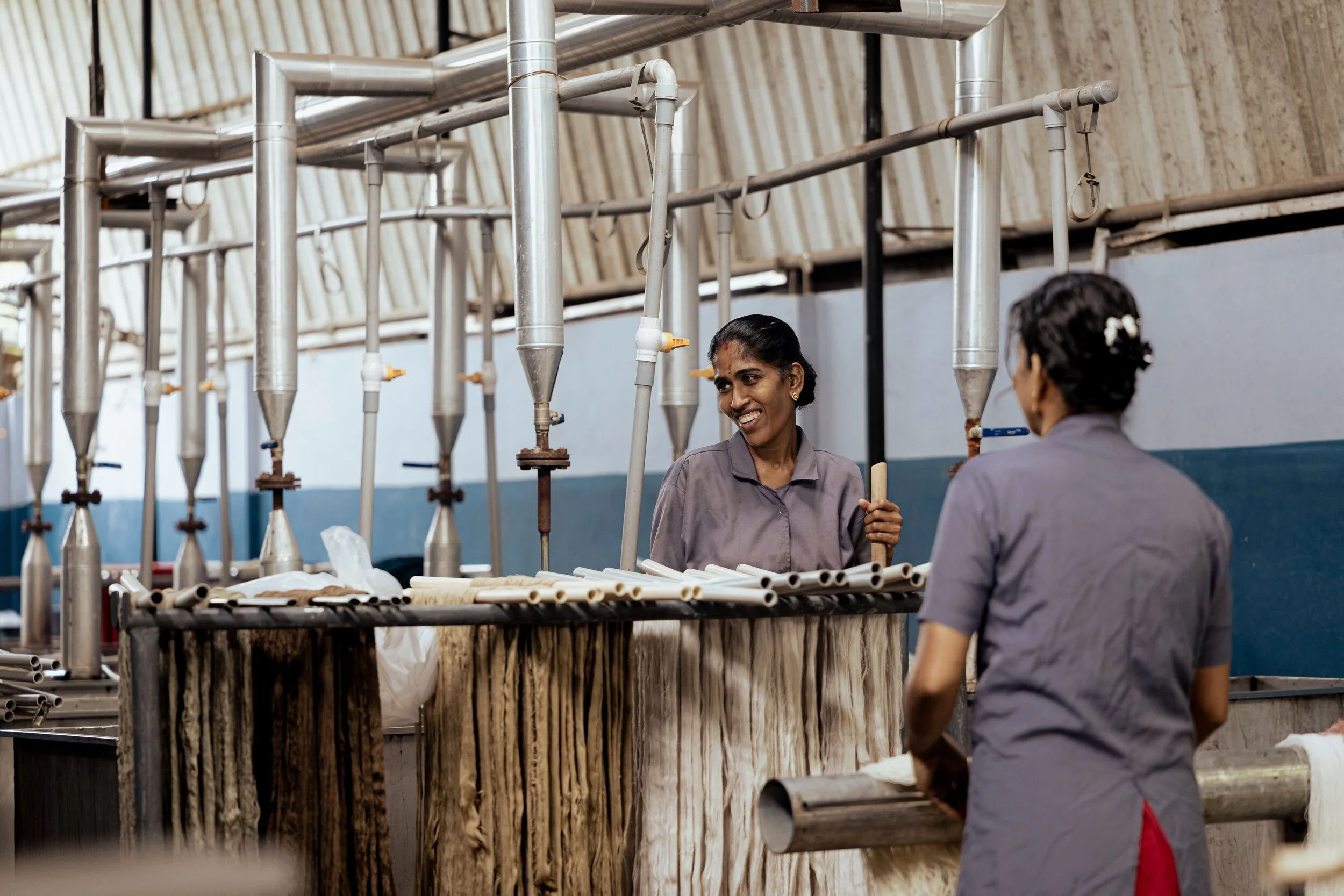 Two women in gray uniforms working with pipes and industrial equipment in a factory or workshop.