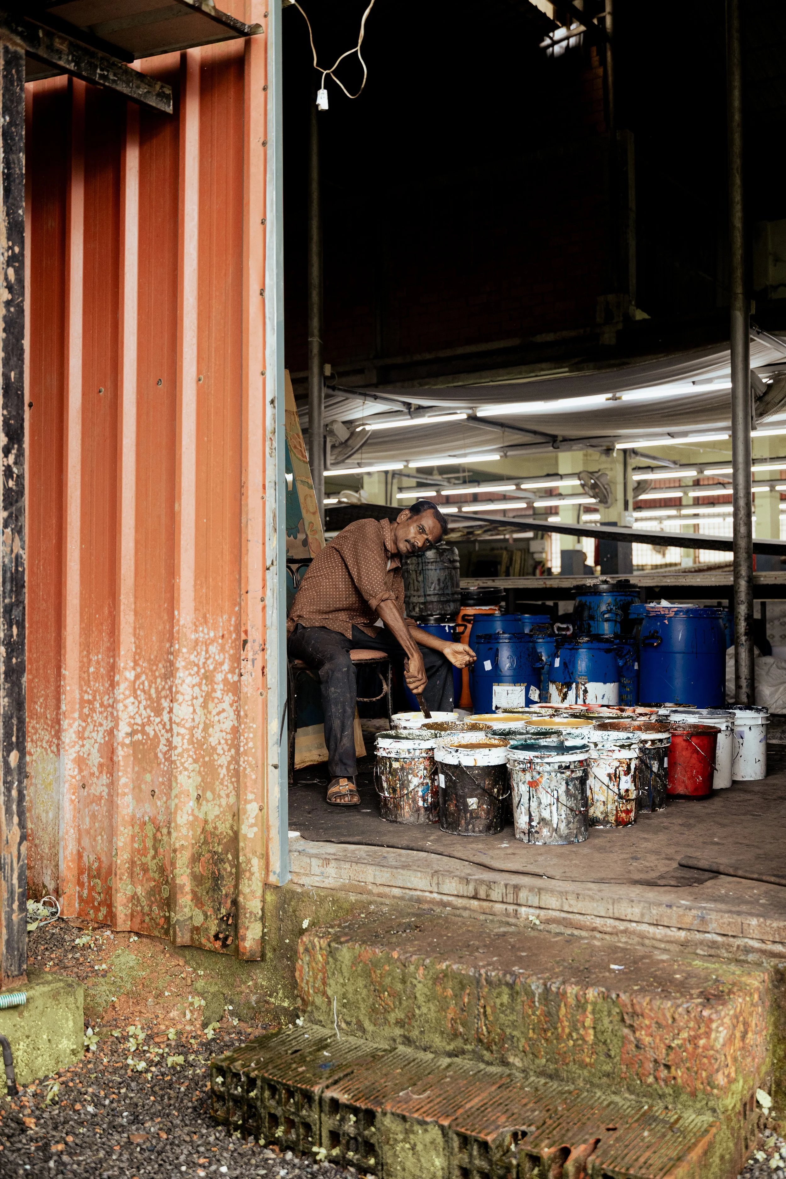 A man sitting on a stool outside a workshop or store, surrounded by large paint buckets with paint splatters, under a roof with fluorescent lighting, with some scaffolding and brickwork visible in the background.