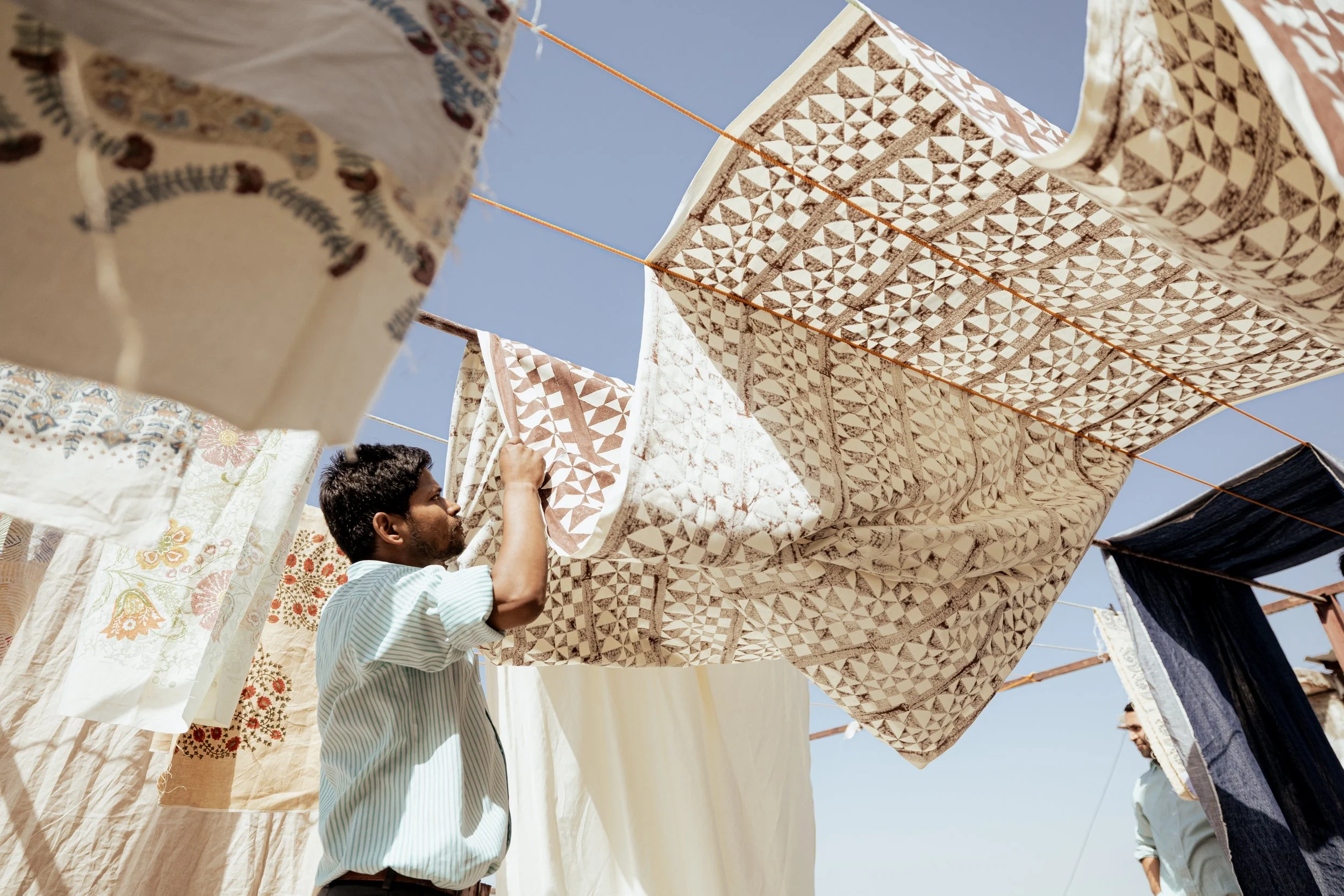 A man hangs patterned fabrics and bed linens on clotheslines outdoors against a clear blue sky, likely drying them in the sun.