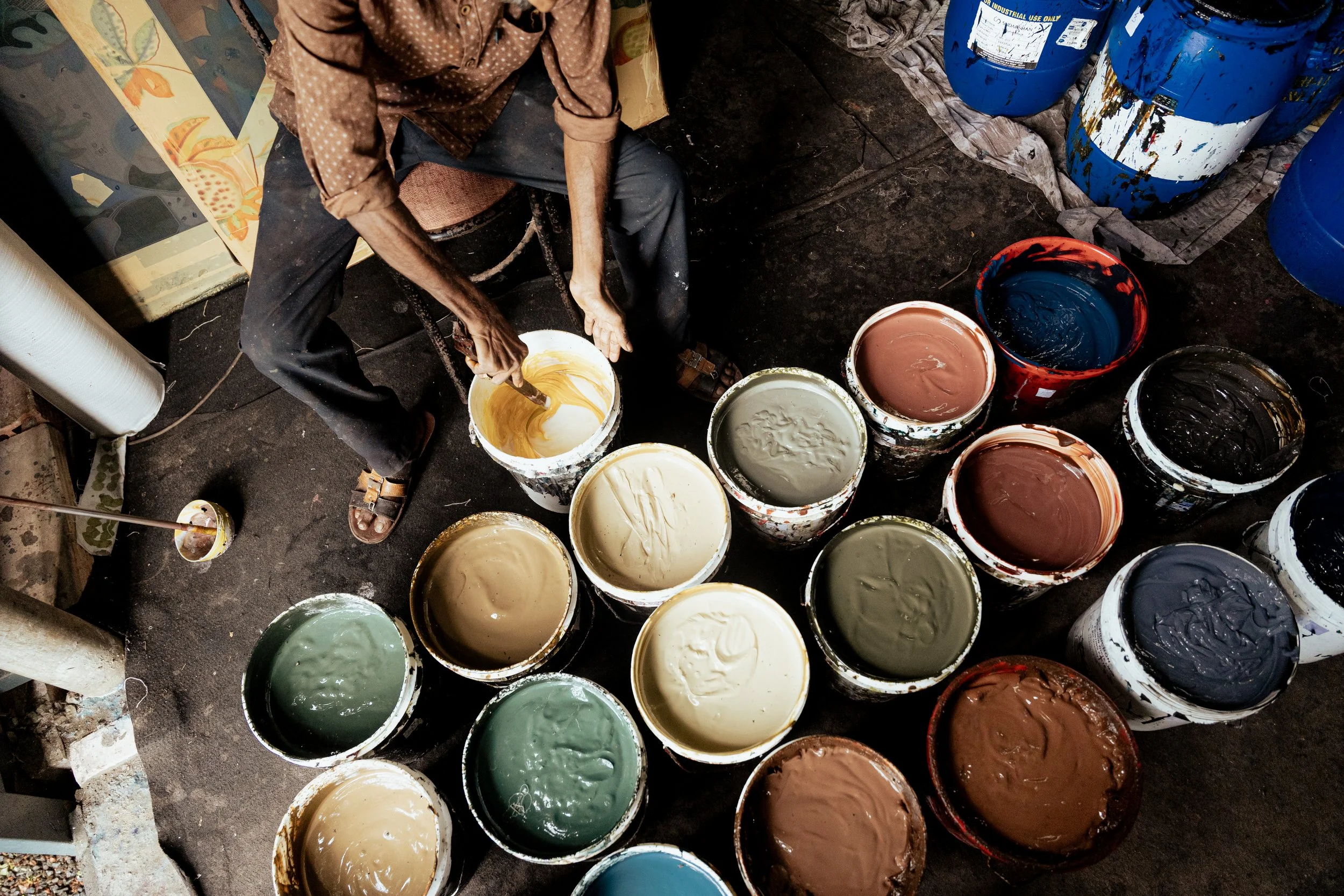 A person mixing paint in several open buckets filled with different colors, along with blue paint buckets and supplies on the floor.