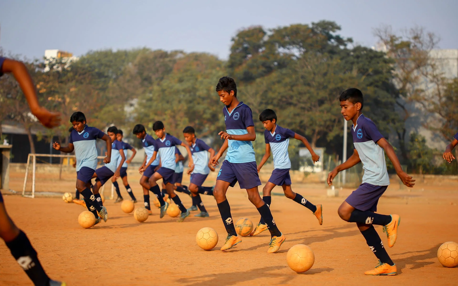Young boys in soccer uniforms practicing dribbling soccer balls on a dirt field, with trees and a goalpost in the background.