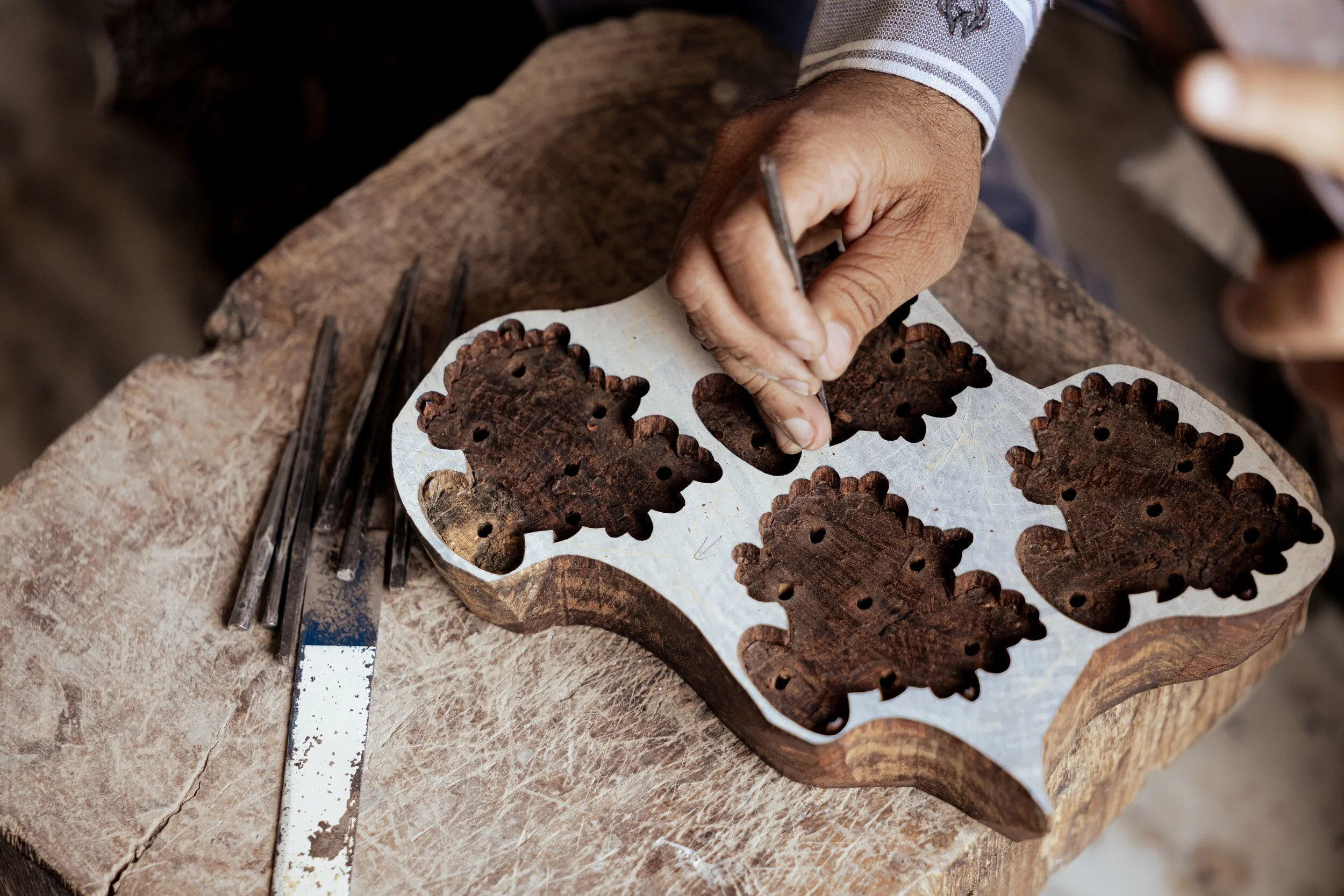 A craftsman working on carved wooden pieces, possibly traditional patterns, on a wooden workbench. The craftsman is using a small tool for detailed carving.