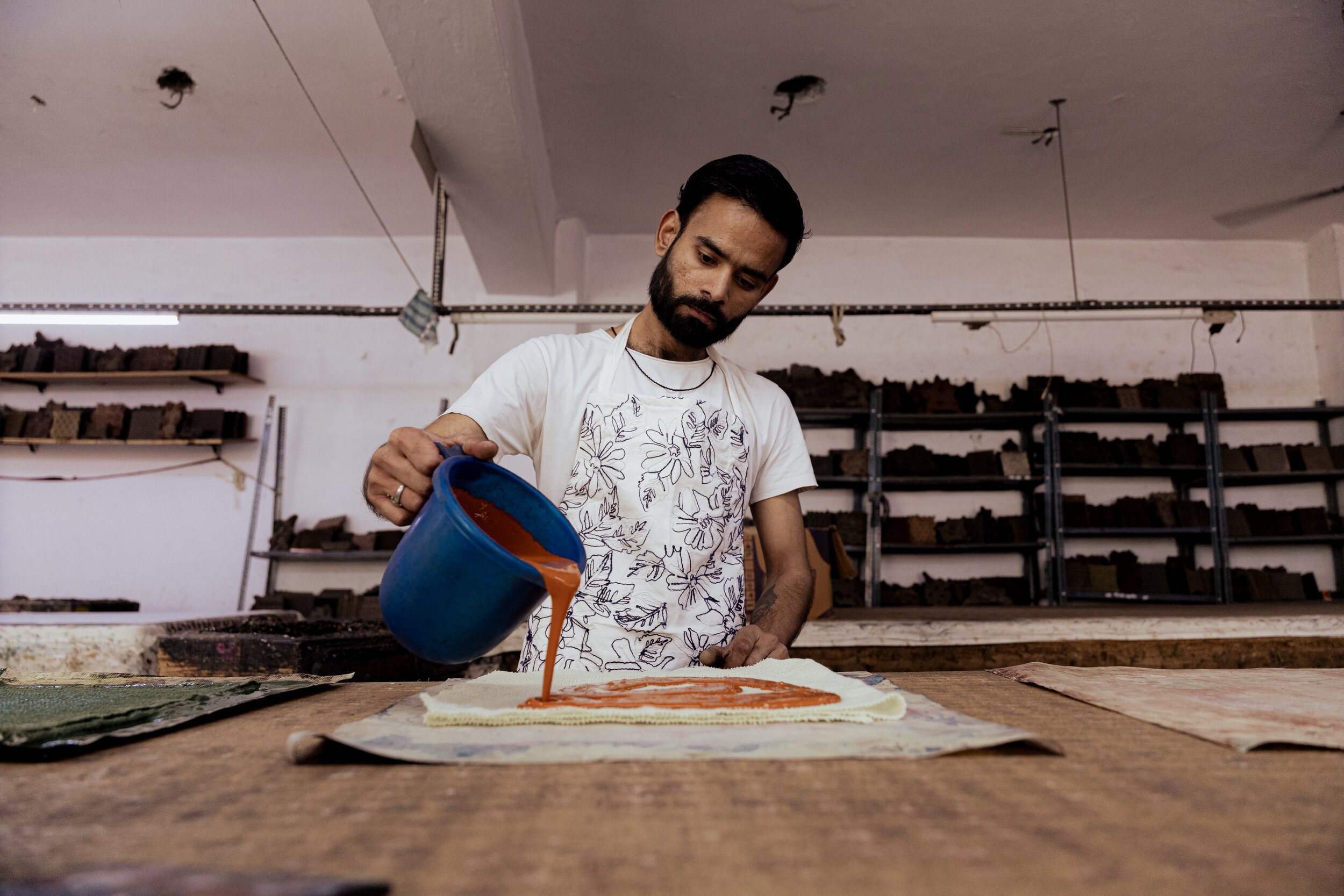 A man with a beard and dark hair pouring orange paint from a blue container onto a piece of fabric or paper on a worktable in an art studio or workshop, with shelves of supplies in the background.