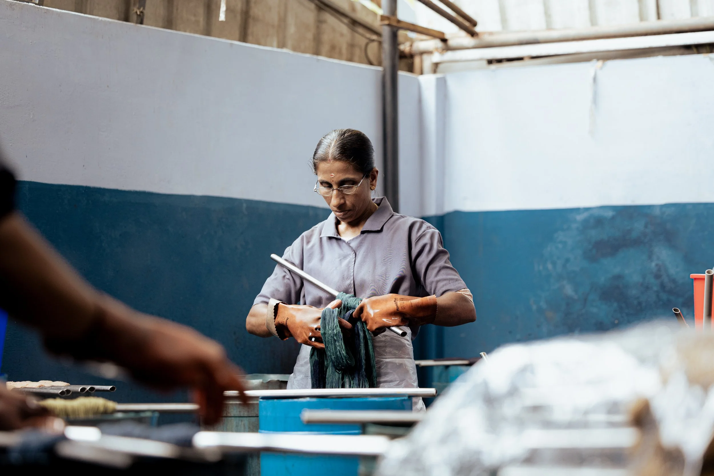 A woman with glasses and traditional attire working with yarn or fabric in an industrial or workshop setting.