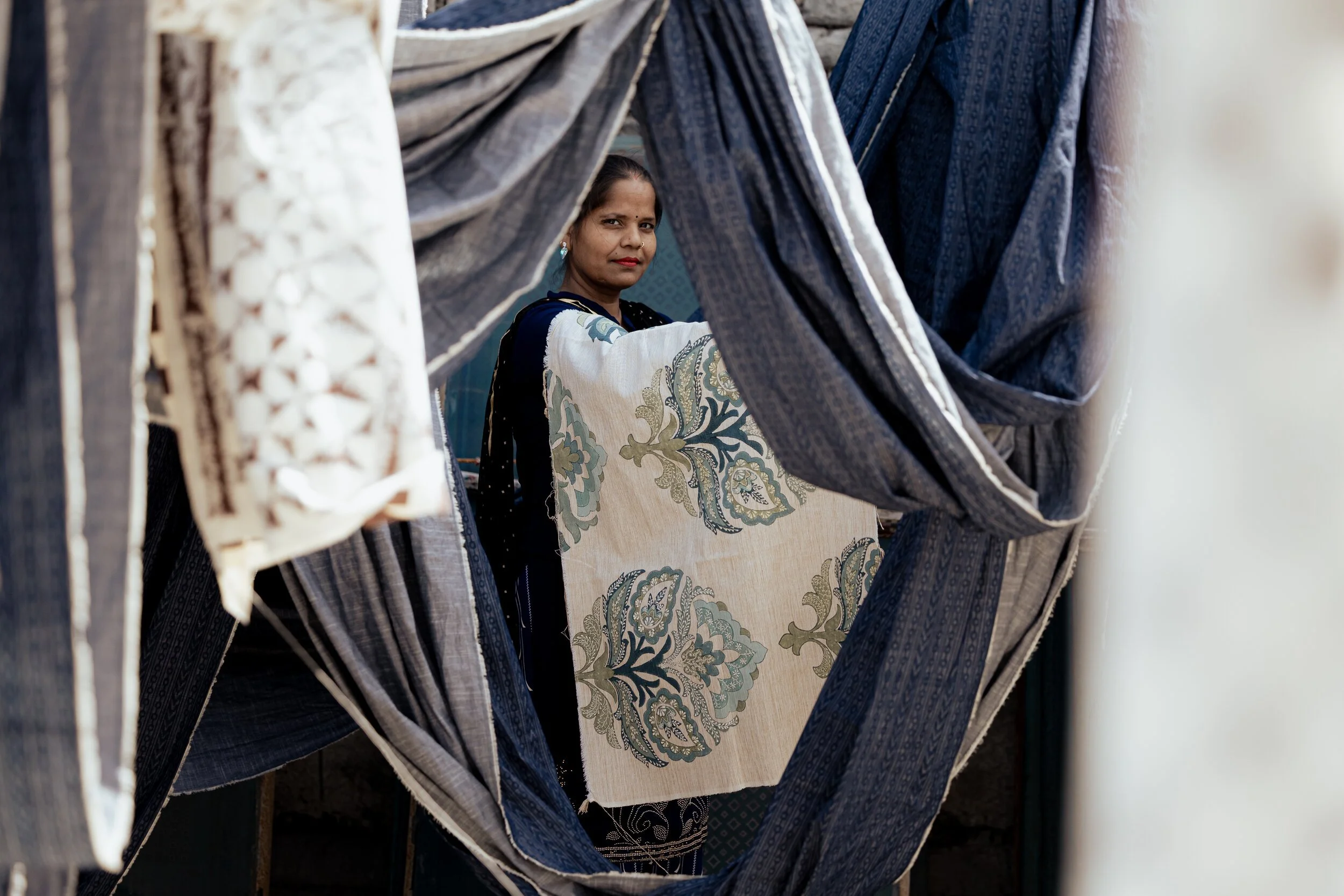 A woman in traditional clothing seen through hanging fabrics with various patterns, holding a large piece of textile with intricate floral designs.
