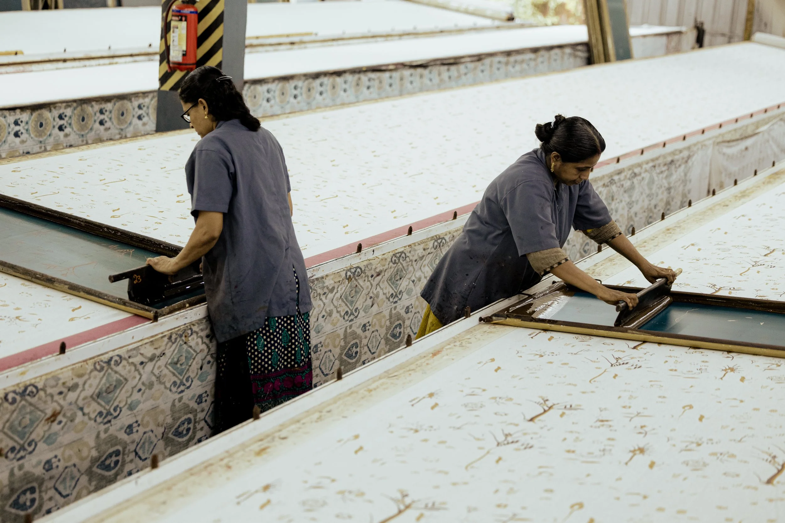 Two women work on a large tile with gold accents in a factory or workshop, wearing dark gray uniforms.