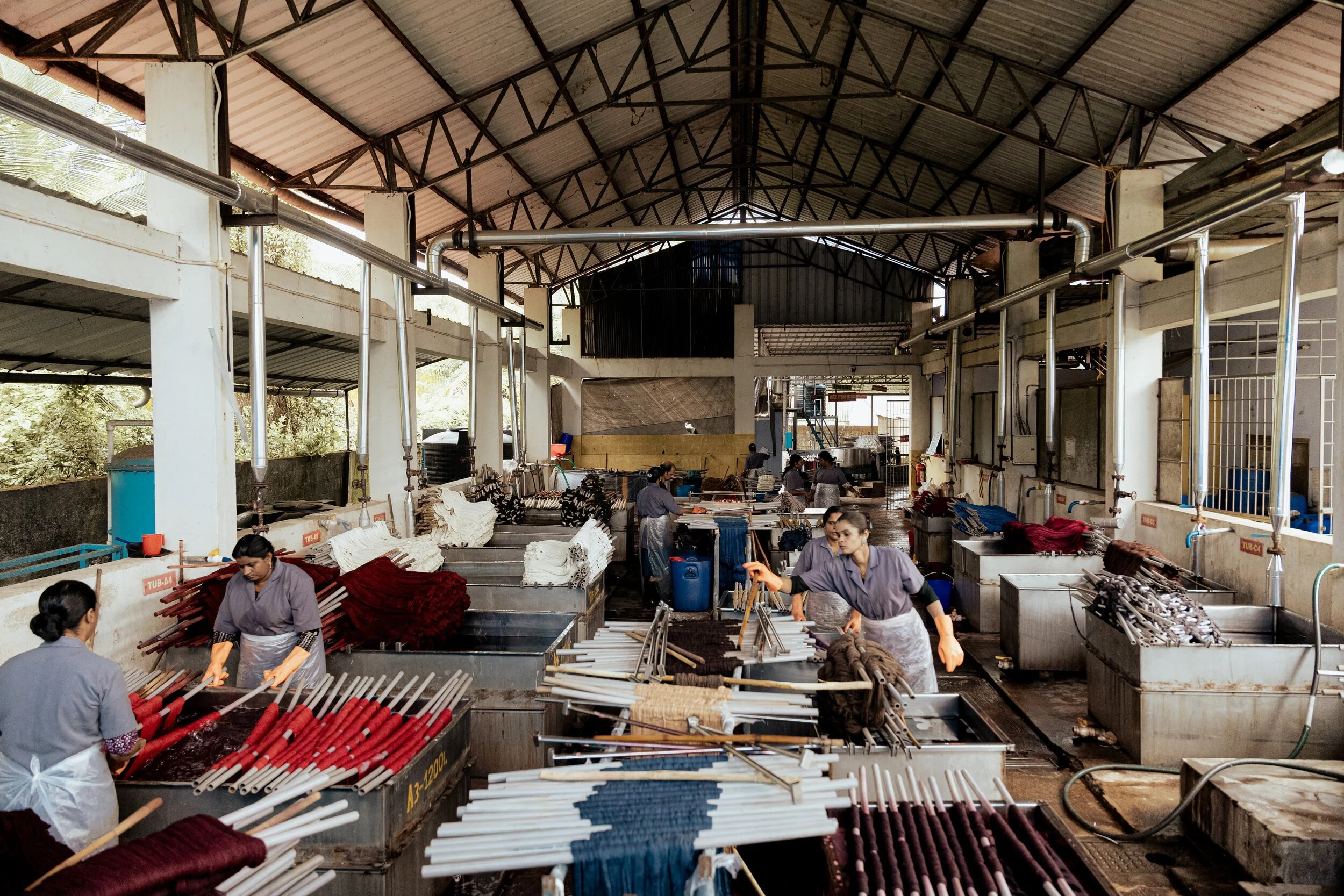 Workers in a factory processing long red and white fabric or foam tubes, with industrial metal pipes and equipment overhead.