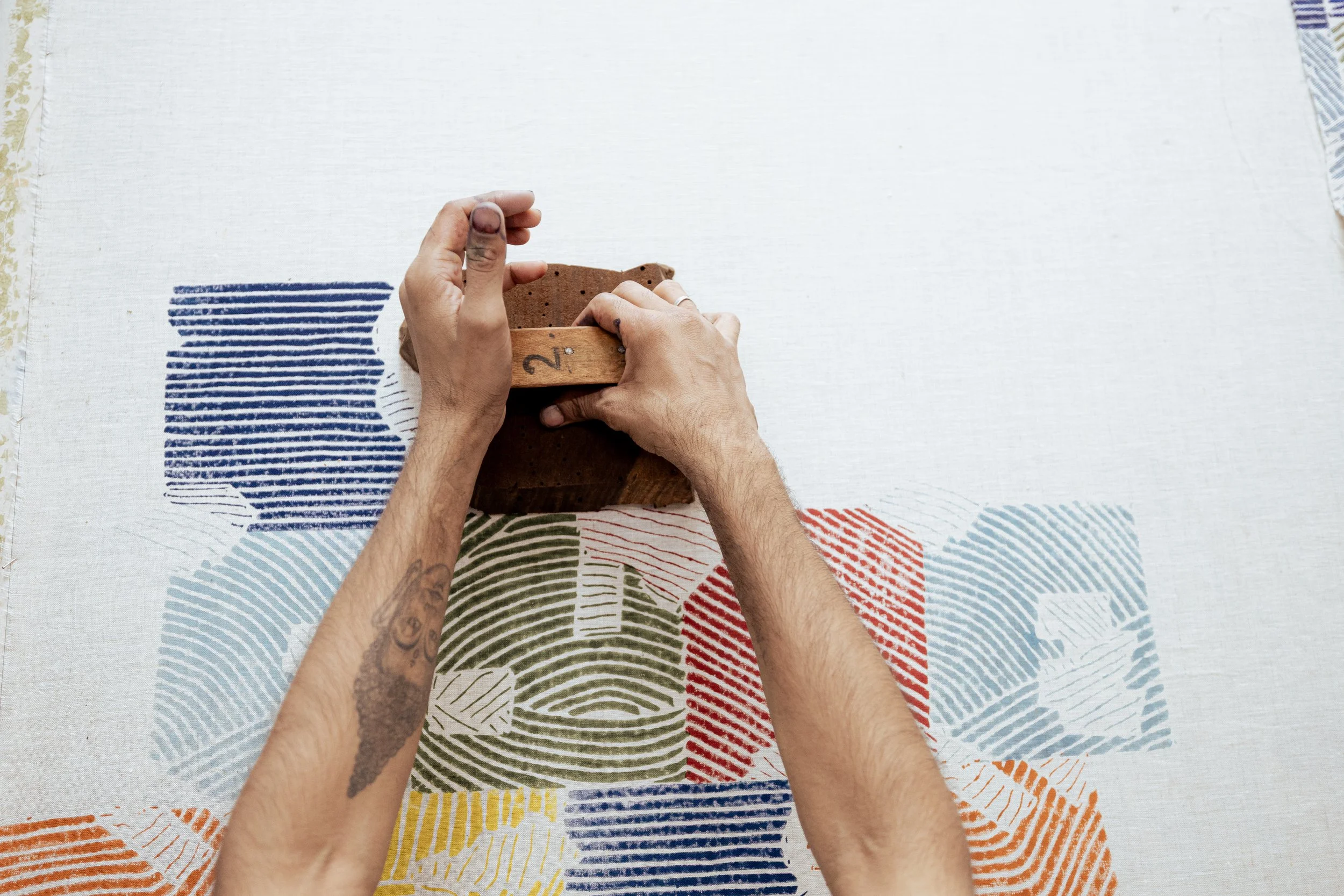 Two hands with tattoos holding a small terracotta flowerpot on a colorful, patterned tablecloth.