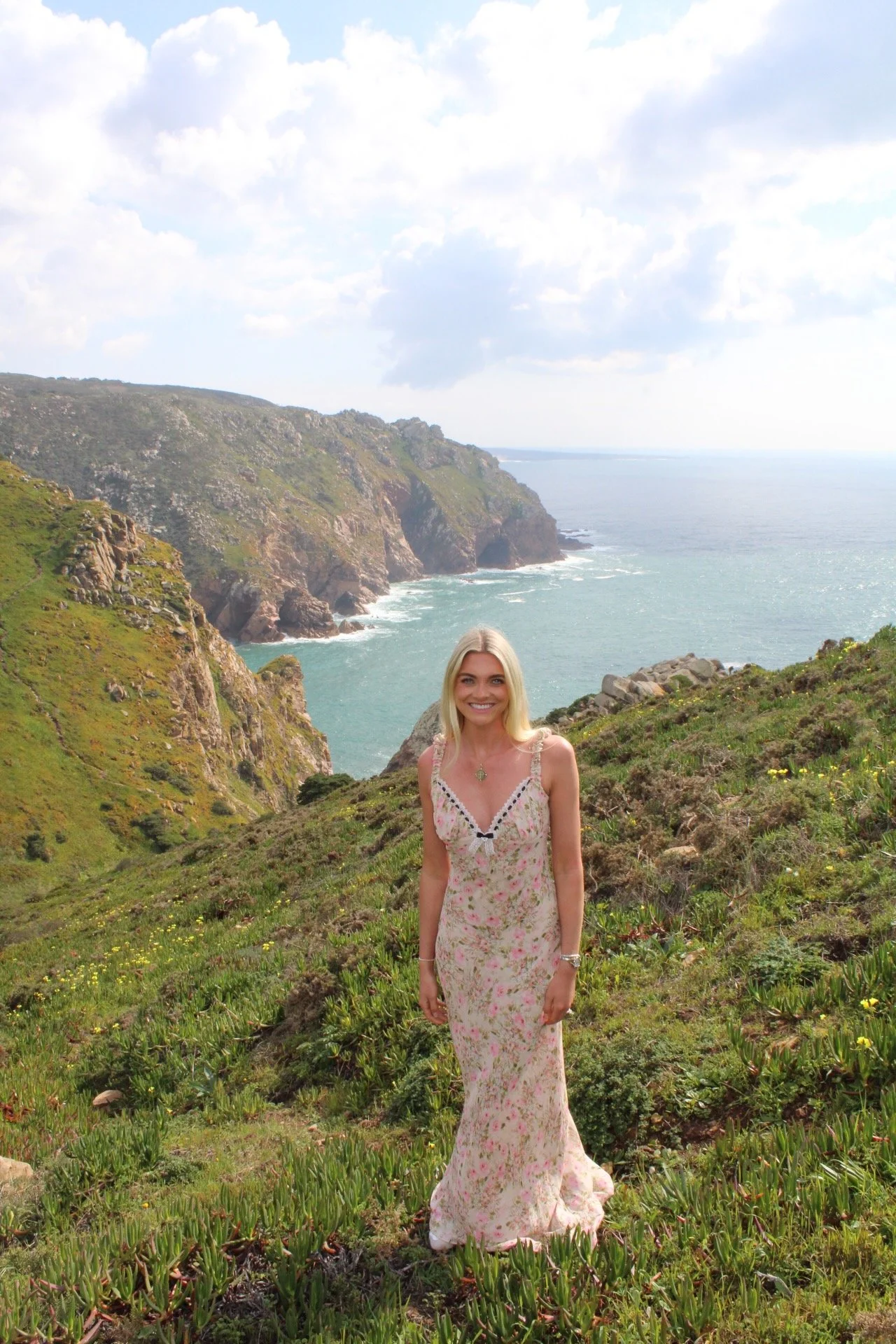 A woman with blonde hair smiling in a long floral dress standing on a grassy hillside with cliffs and ocean in the background.