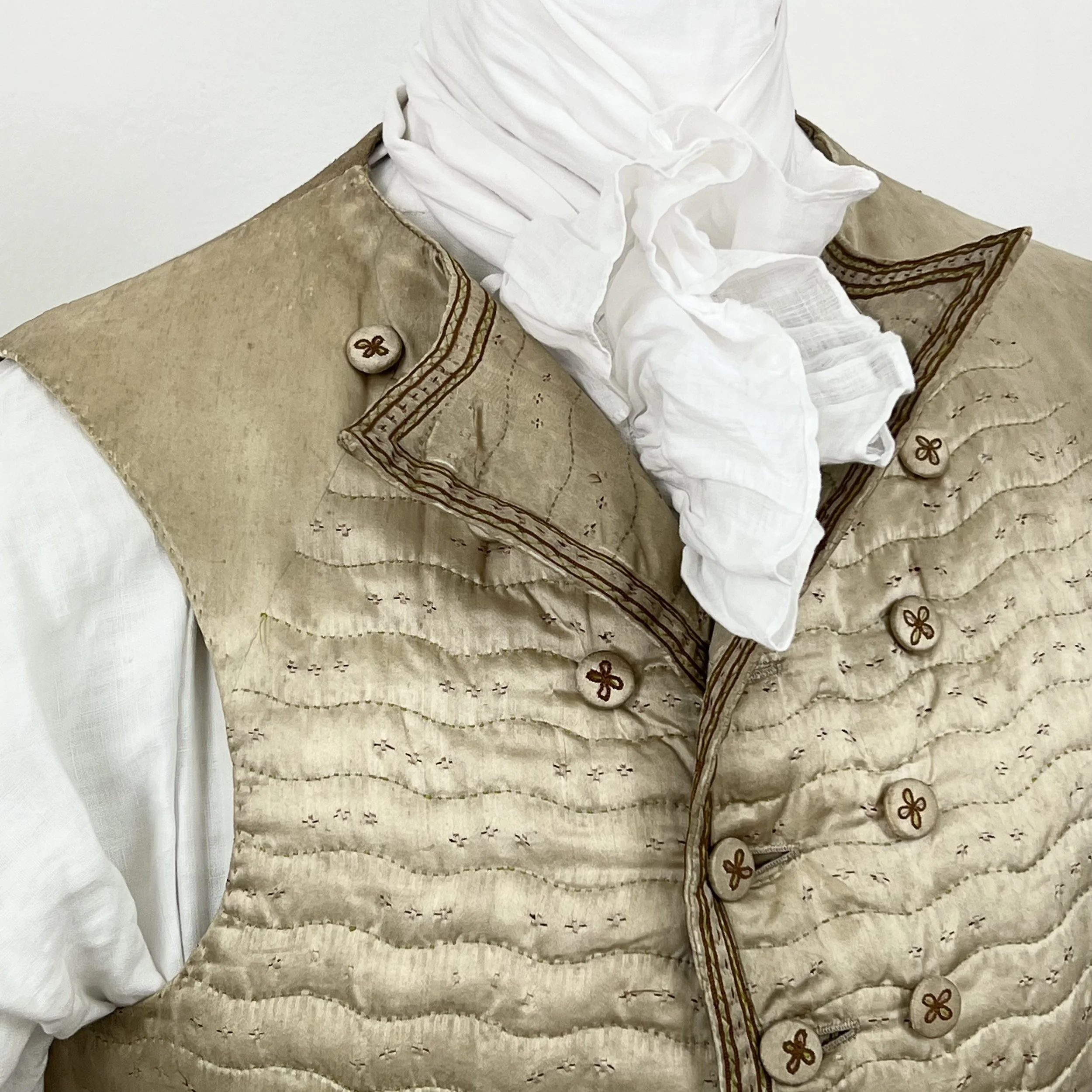 Close-up of a beige quilted vest with floral buttons and embroidered details, worn over a white shirt with a ruffled white necktie.