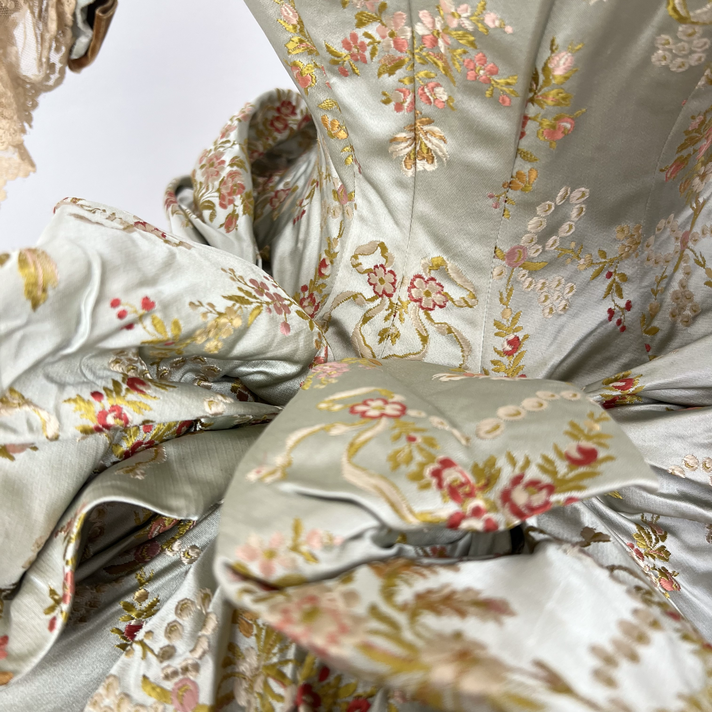 Close-up of a silky, embroidered fabric with floral patterns in pink, gold, and green, featuring intricate detailing and swirling designs.