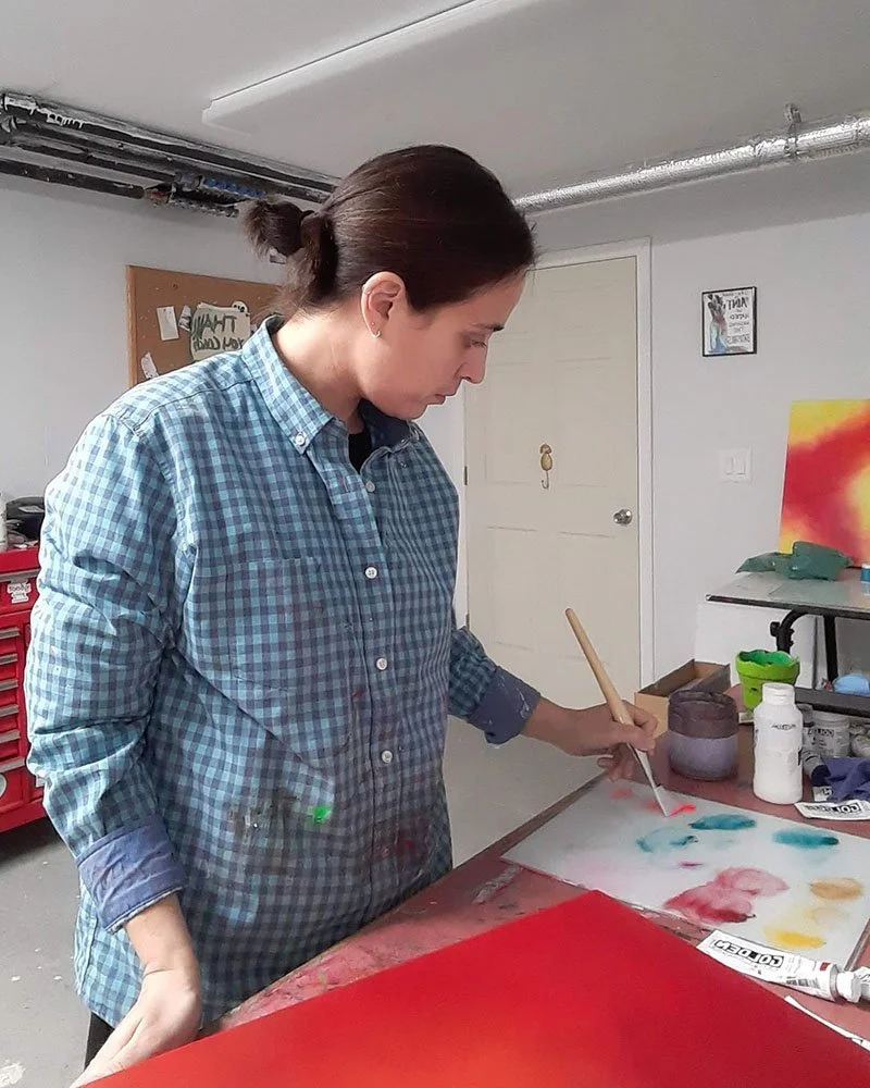 Artist Jennifer Morris working in her studio, holding a paintbrush over her palette with paint-splattered shirt, surrounded by abstract paintings, paint supplies, and studio equipment