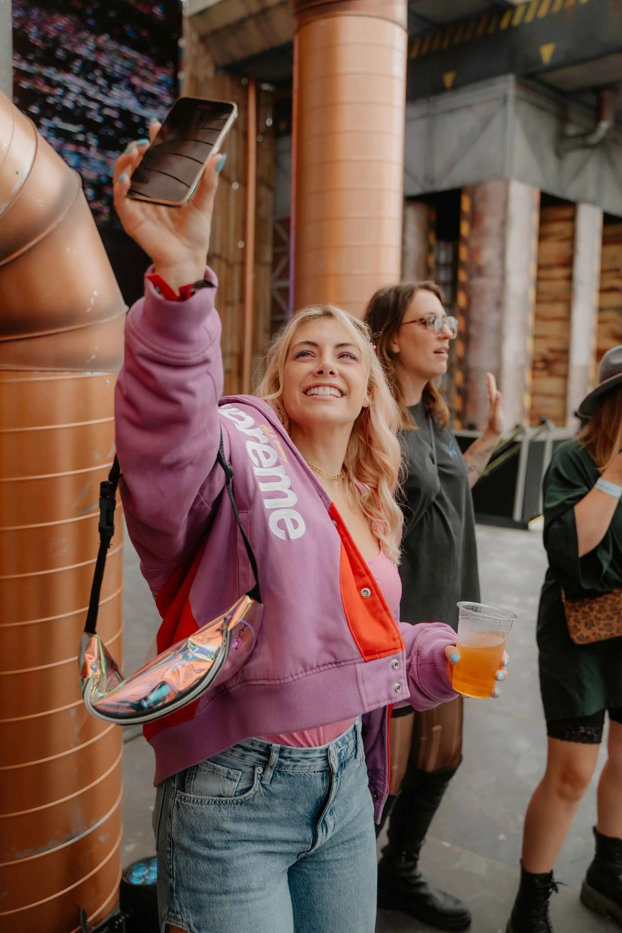 Eine lachende junge Frau mit blonden Haaren, in rosa Mantel und blauen Jeans, macht ein Selfie in einem Veranstaltungsort mit einer metallischen und Holz-Umgebung. Sie hält ein Getränk in der Hand.