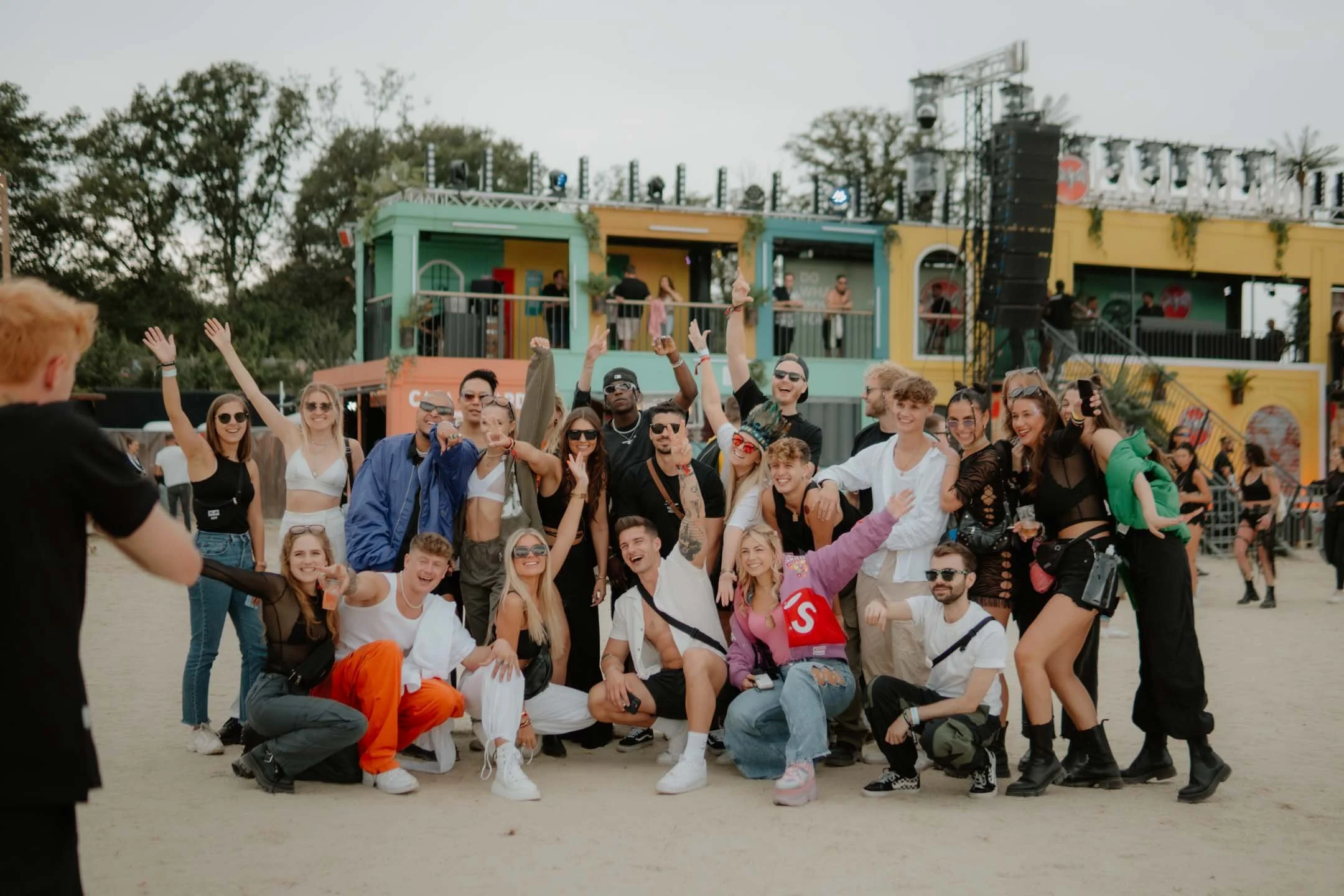 Eine Gruppe junger Menschen posiert auf einem Festivalgelände mit bunten Gebäuden im Hintergrund, einige winken, lachen und feiern.