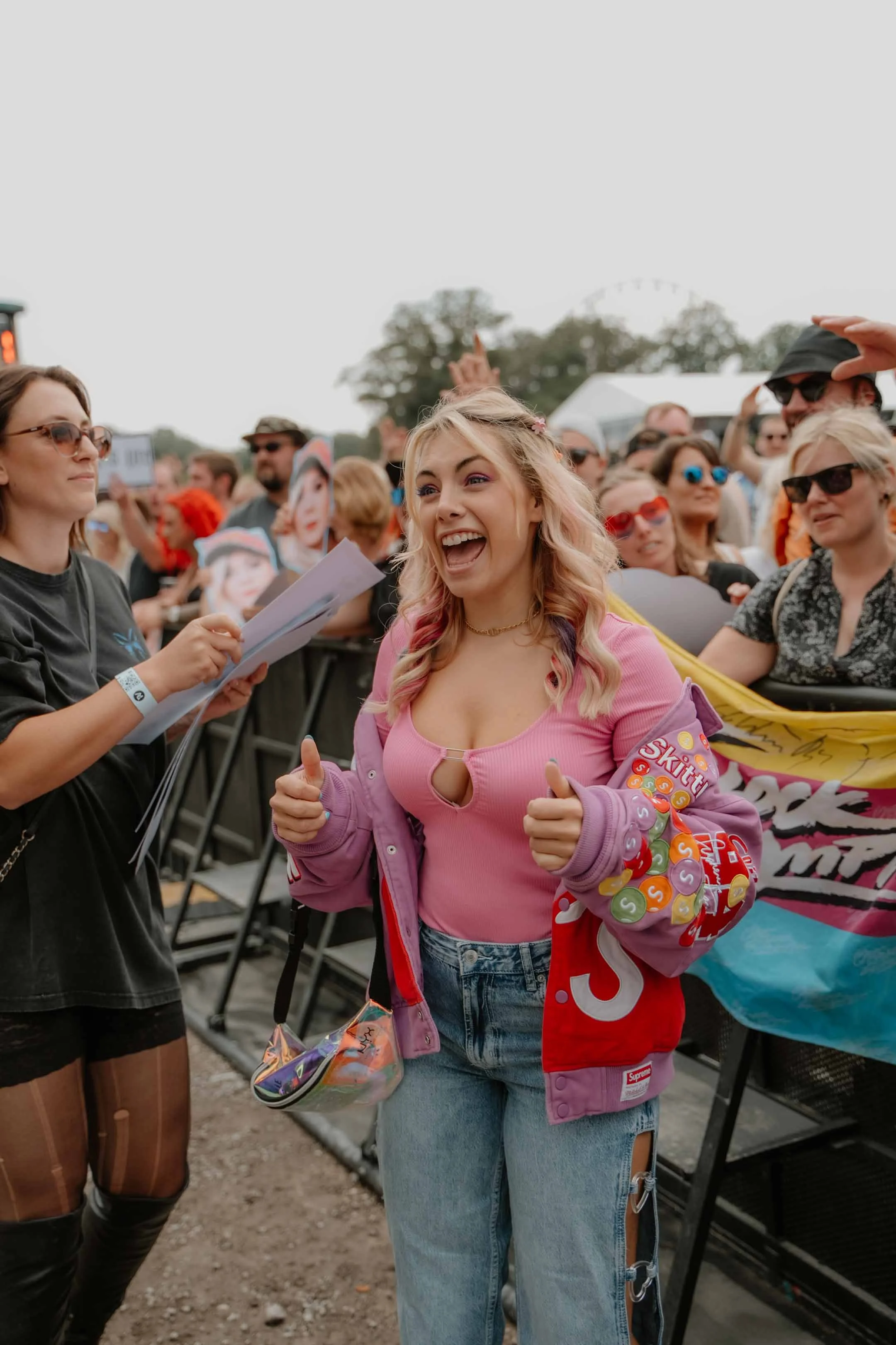 Eine lachende junge Frau beim Festival, umgeben von anderen Festivalbesuchern mit bunten Sonnenbrillen und Bannern im Hintergrund.