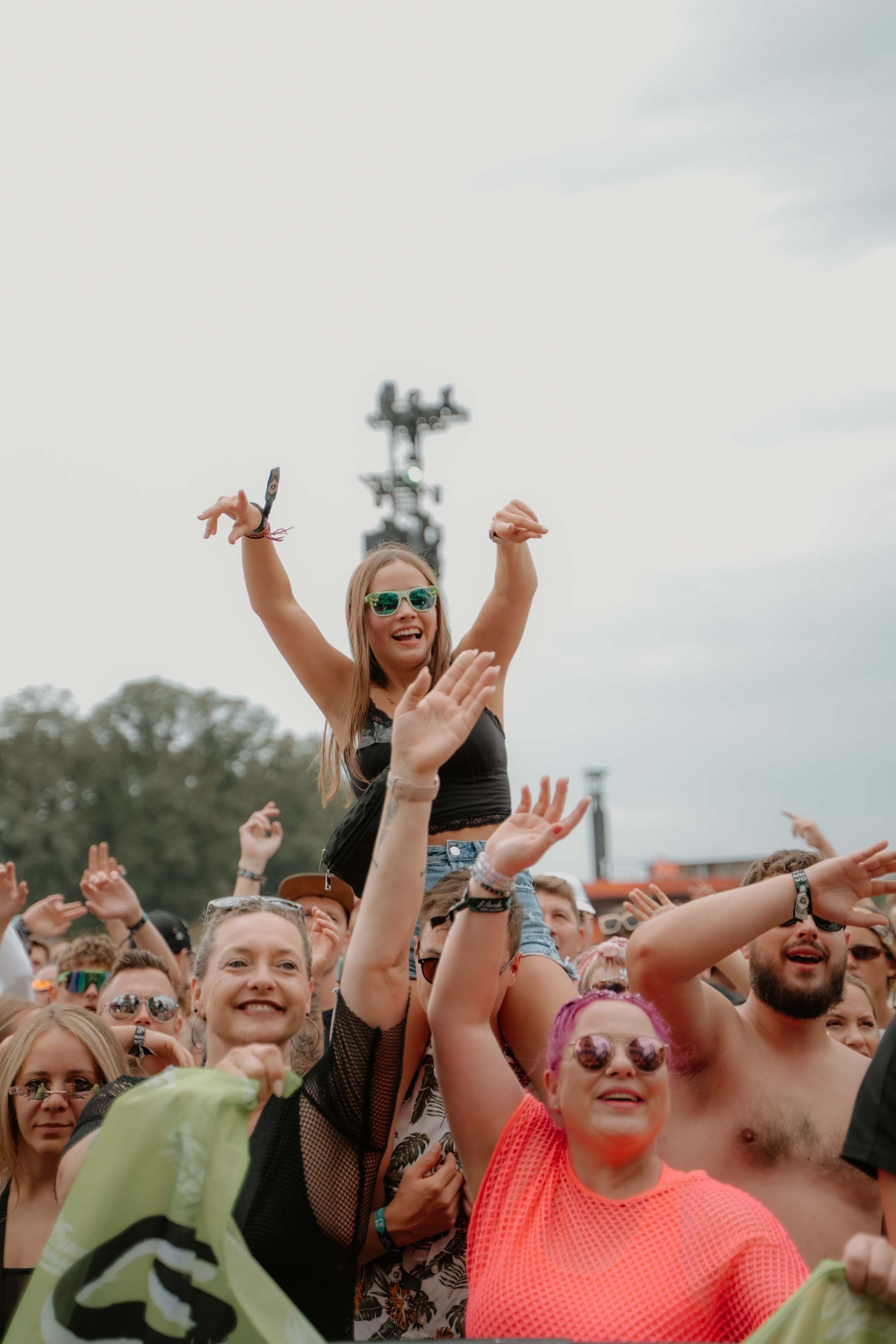 Menschen bei einem Musik- oder Festivalevent, eine Frau sitzt auf den Schultern eines Mannes, trägt Sonnenbrille und lacht, umgeben von weiteren feiernden Personen.