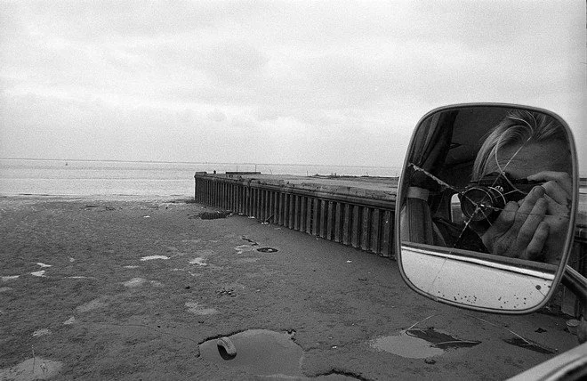 A black and white photo of a beach scene with a breakwater extending into the water. The image is reflected in the side mirror of a vehicle, showing a person taking the photo. The ground is sandy with small puddles and scattered debris.