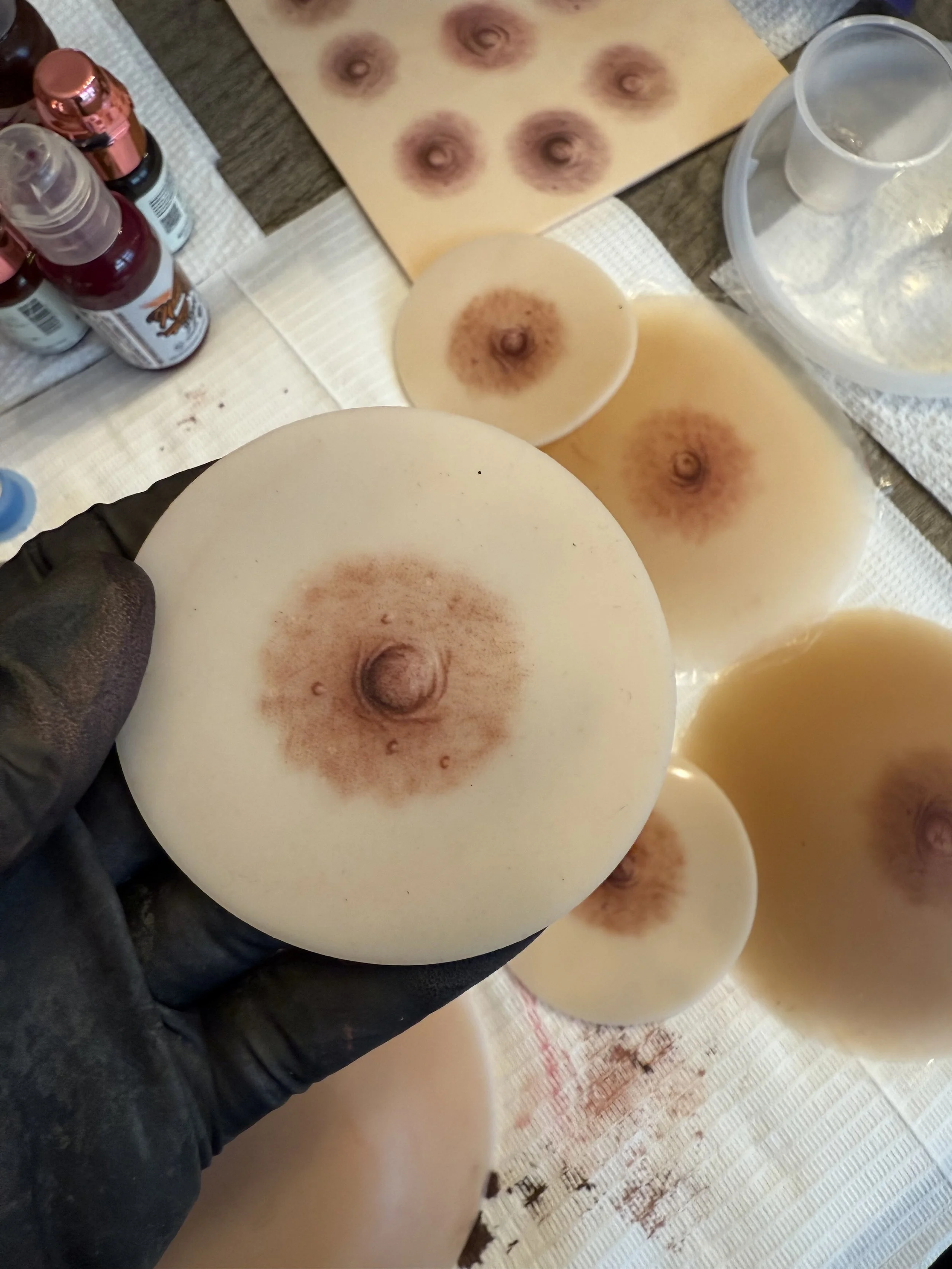 Close-up of a person's hand in a black glove holding a circular object with a hole in the center and brown markings, on a table with several similar objects and various bottles, paper towels, and a container in the background.