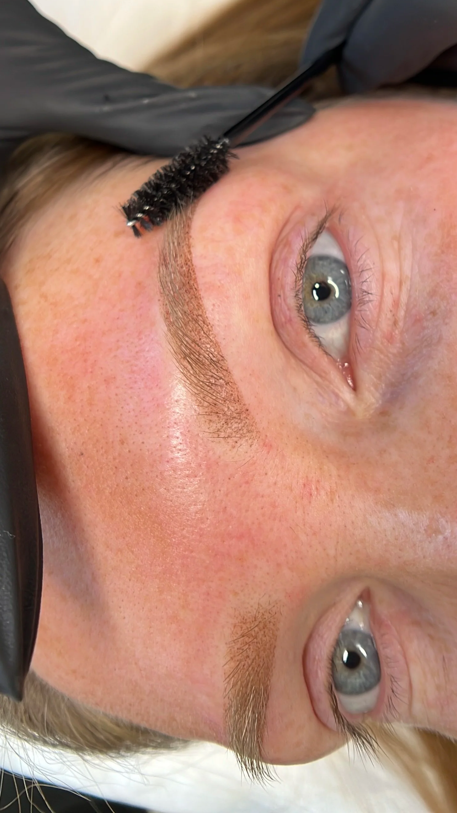 A person lying down with their eye and eyebrow visible, a black brush is applying makeup or cosmetic product to the eyebrow.