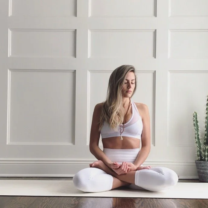 A woman practicing yoga indoors, sitting cross-legged on a mat with eyes closed in a meditative pose.