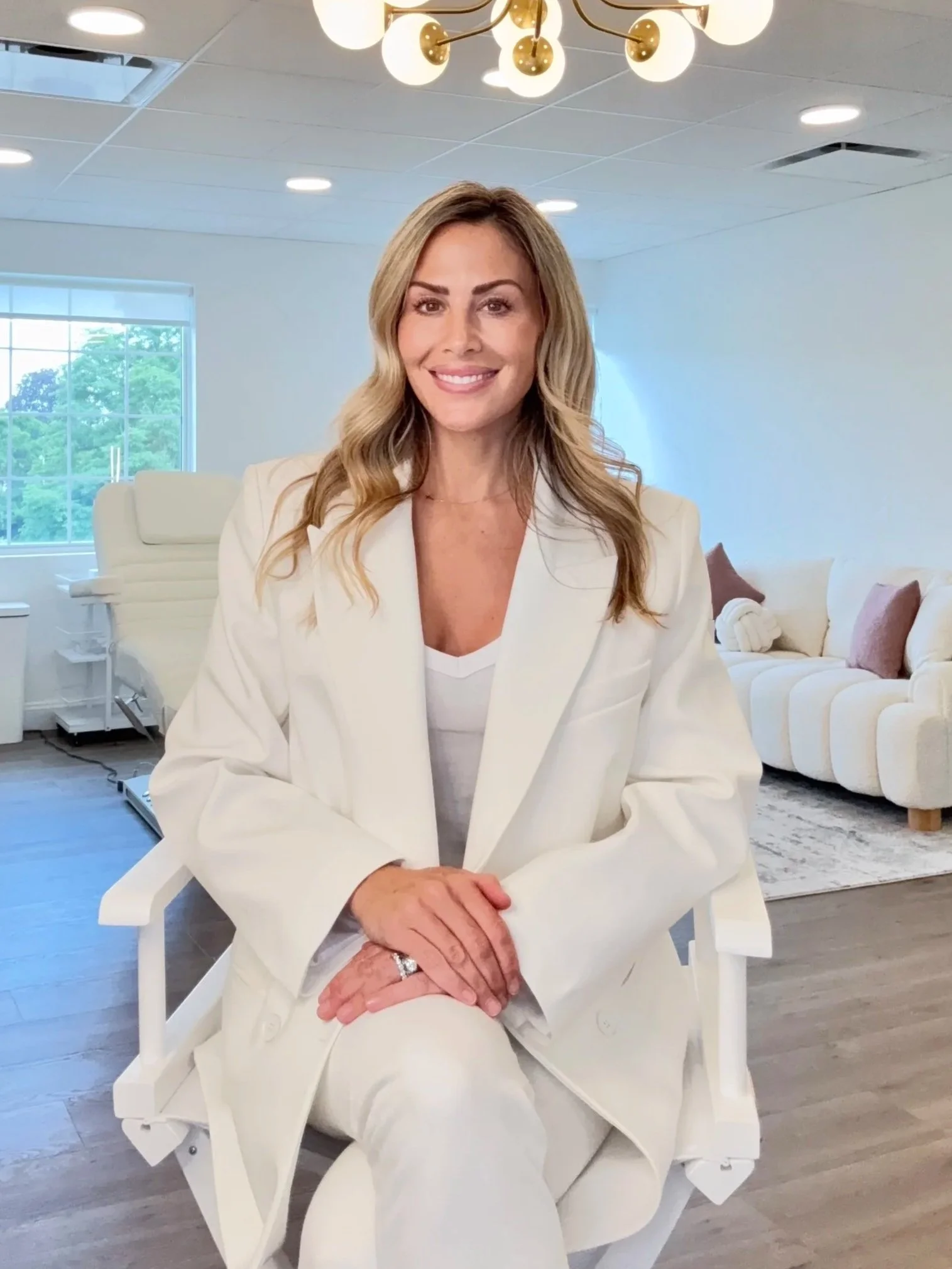 A woman with shoulder-length blonde hair, wearing a white blazer and white pants, seated in a modern, well-lit room with a white sofa and large window showing outside greenery. She is smiling, with her hands resting on her lap, and a chandelier with spherical lights hangs from the ceiling.