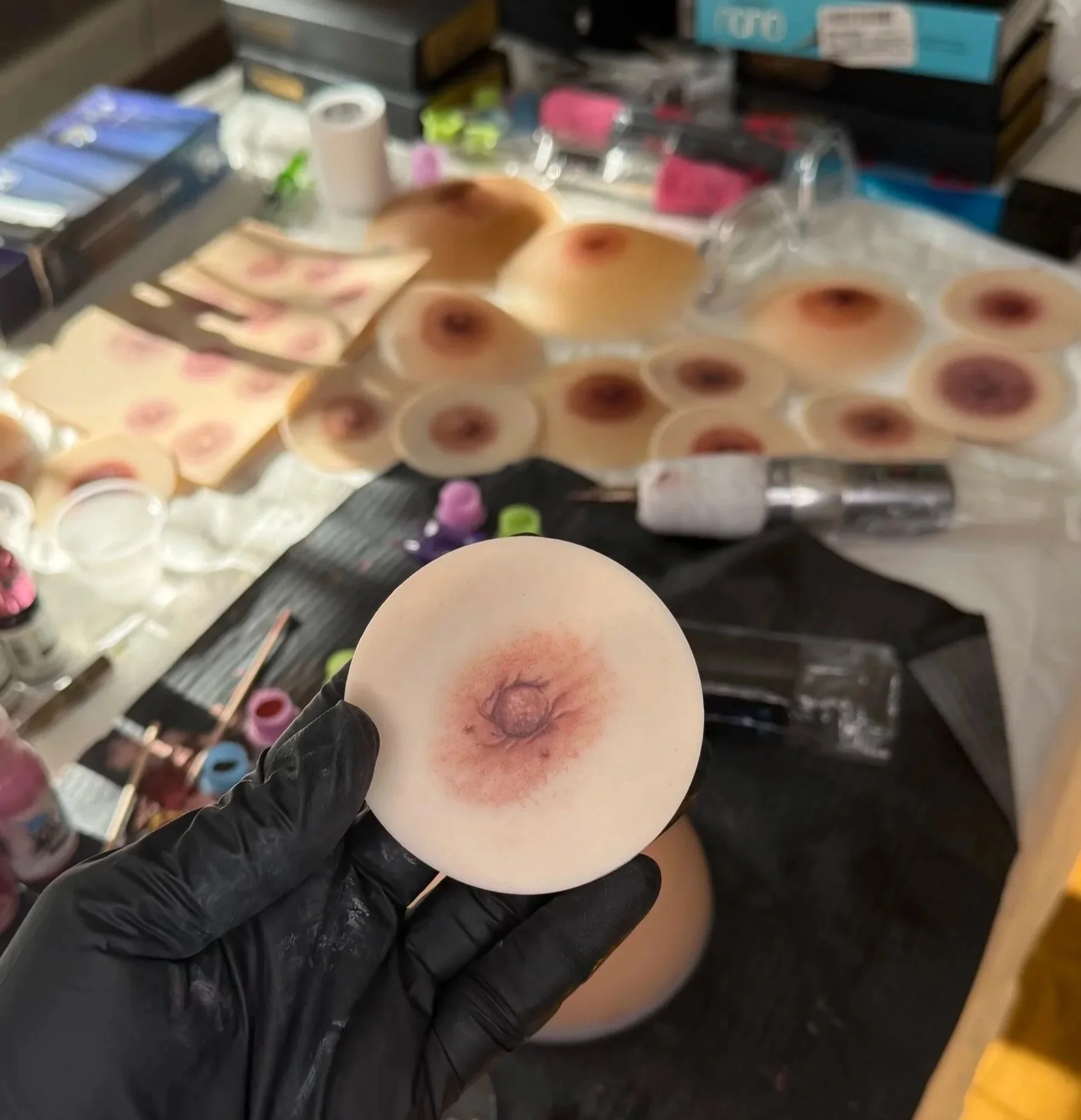A person wearing a black glove holding a raw egg. The egg has some redness and bruising around its yolk. In the background, there are various art supplies and craft items, including boxes, tape, and small containers.