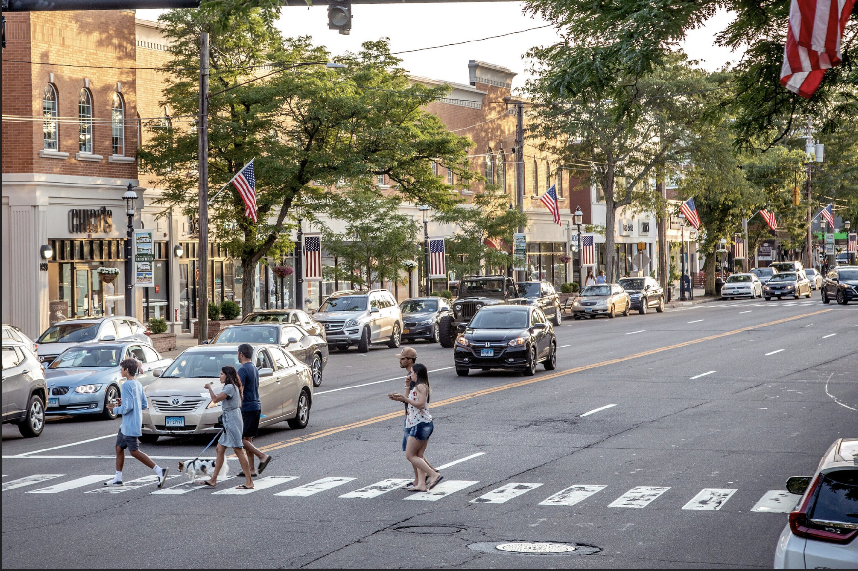 People walking across a crosswalk in a small town street with parked cars and storefronts decorated with American flags and trees.