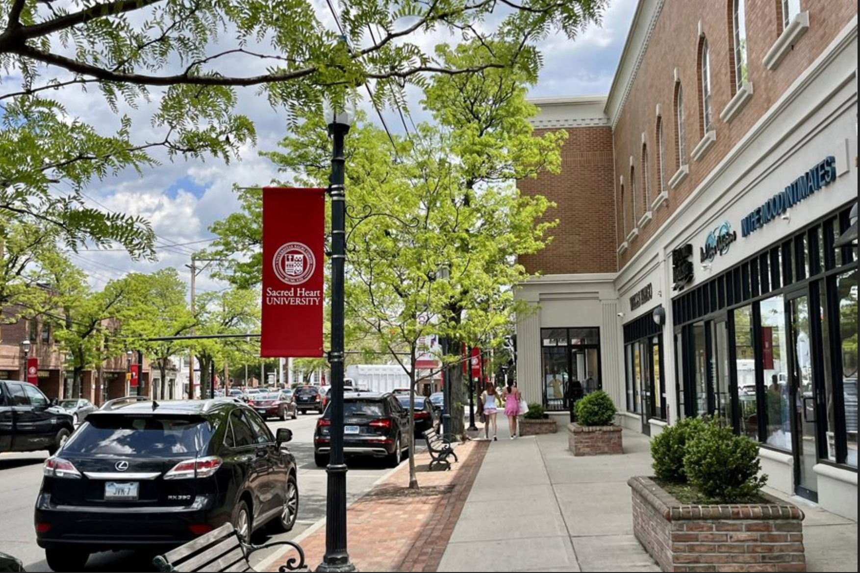 Street view showing a shopping district with a Sacred Heart University banner, trees, parked cars, a sidewalk, and storefronts including a store called Little Moonmenaties.