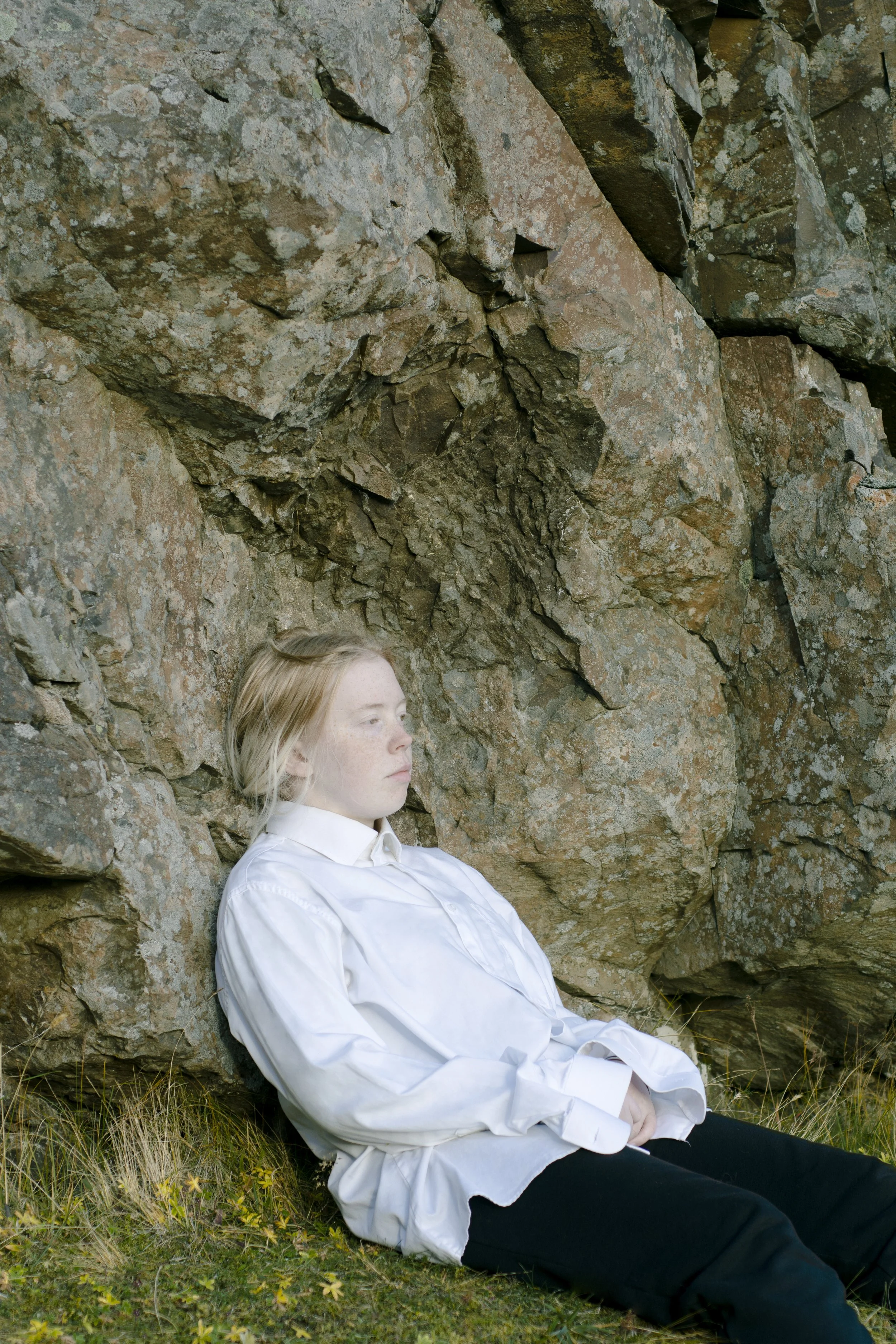 A young person with blonde hair and freckles wearing a white shirt and black pants sitting against a large rocky wall in a natural outdoor setting.