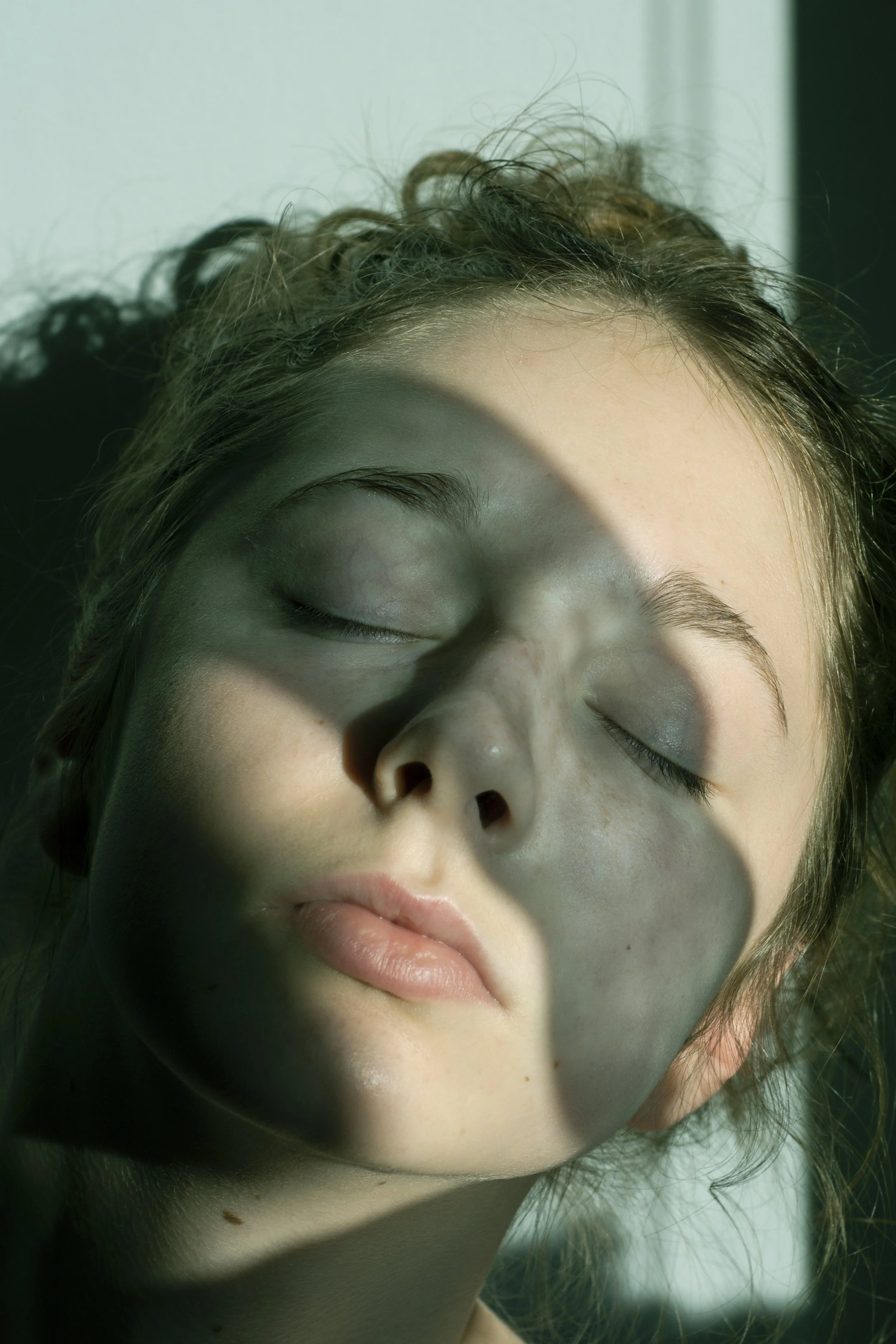 Close-up of a young woman with closed eyes, experiencing sunlight and shadow across her face, with parts of her face in a shadow cast by an object or window.