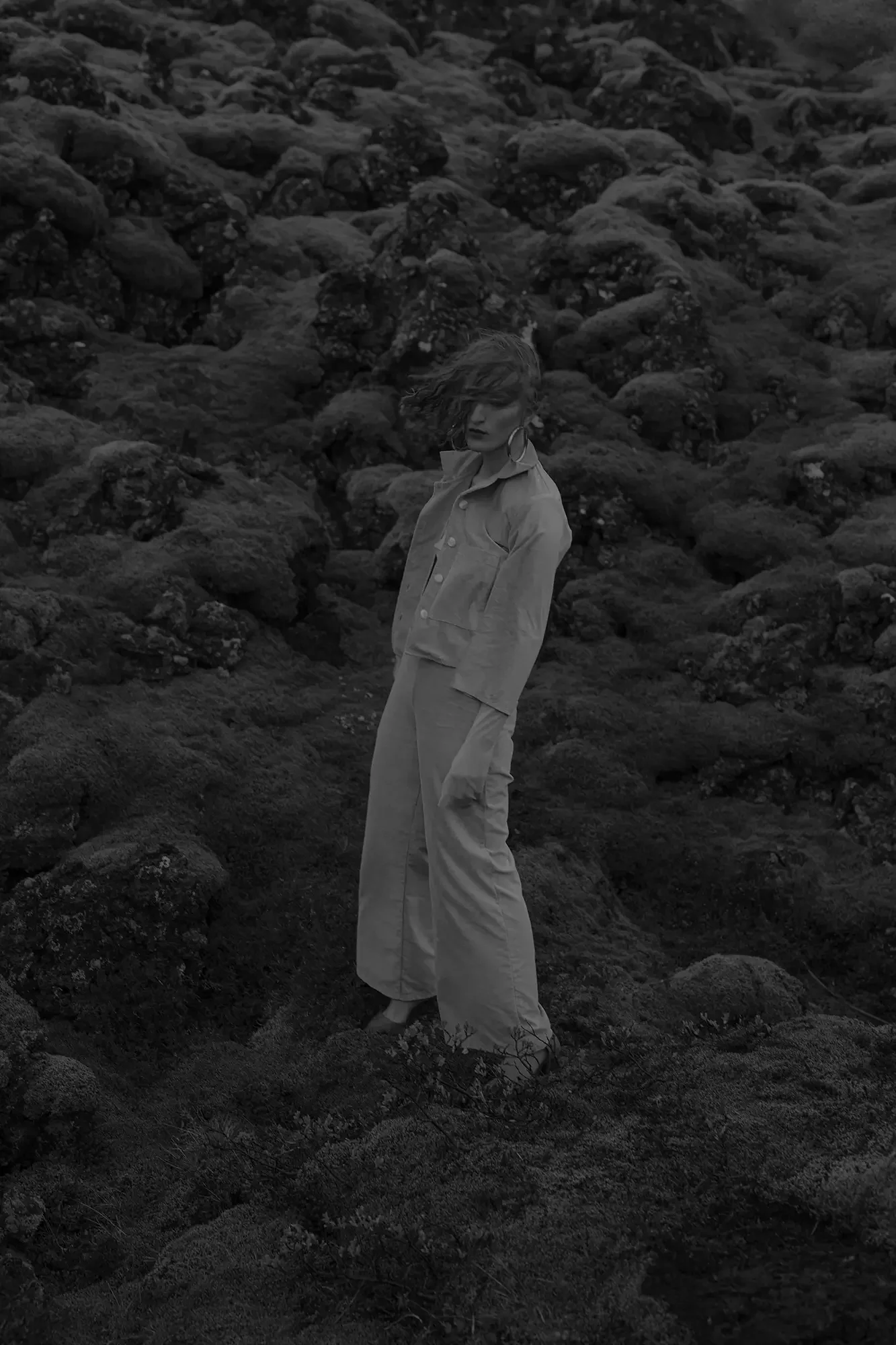 A woman standing on rocky terrain wearing a jacket and wide-leg pants, with wind blowing her hair across her face.