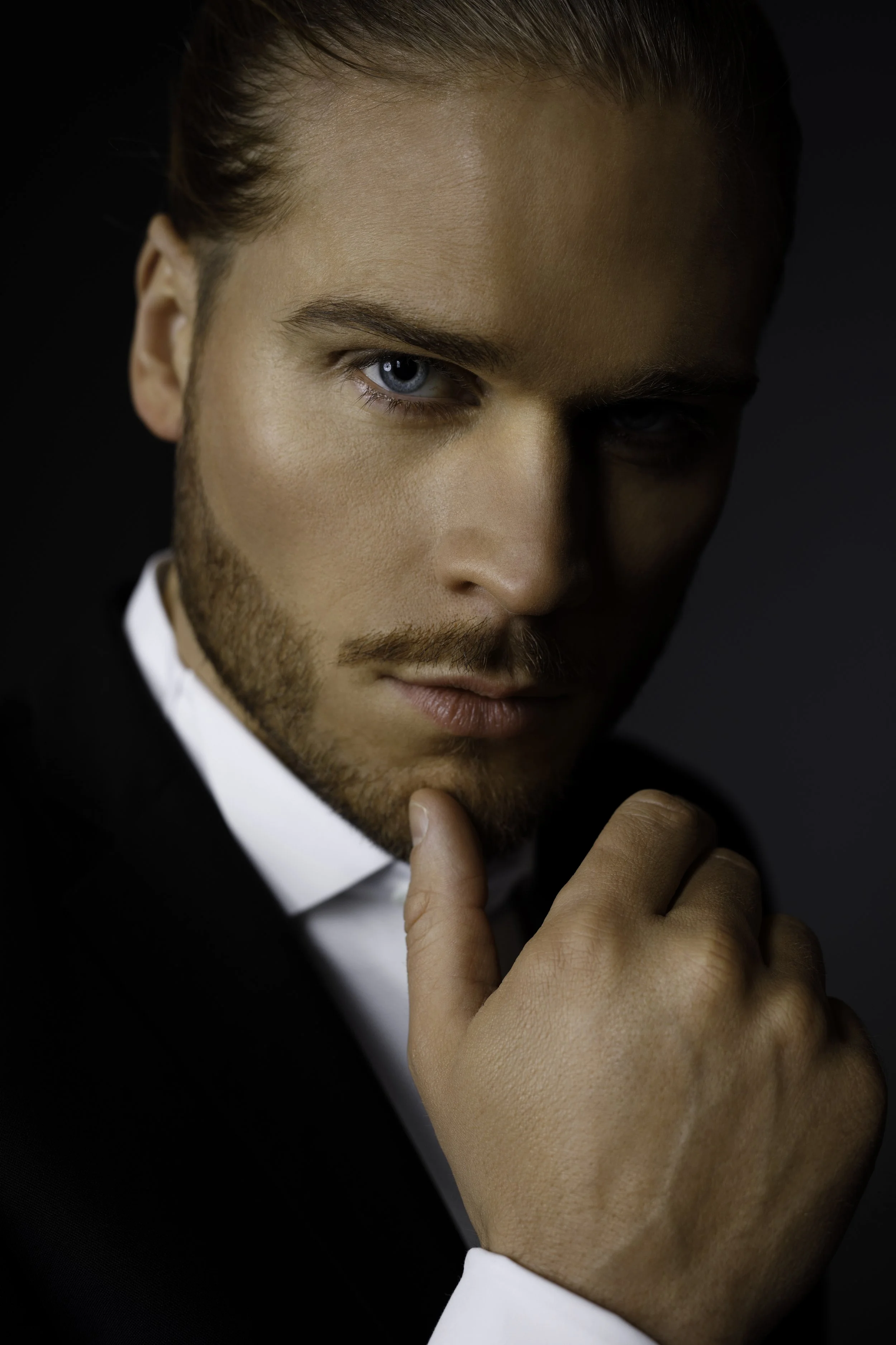 Close-up portrait of a man with blue eyes and light brown hair, wearing a black suit and white shirt, posing with his hand near his chin against a dark background.