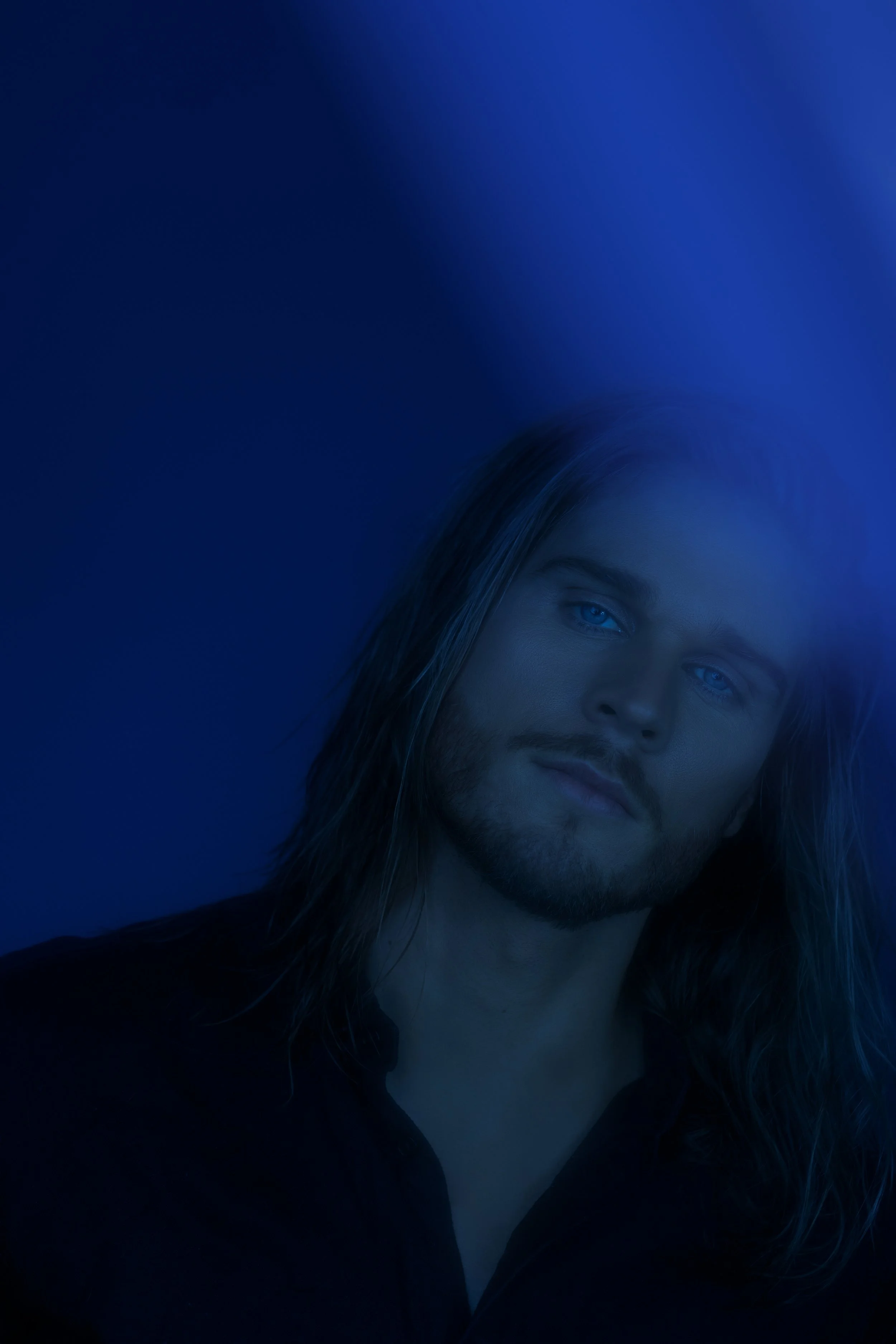 Portrait of a man with long hair and a beard, looking slightly to the side, with a dark background and blue lighting.