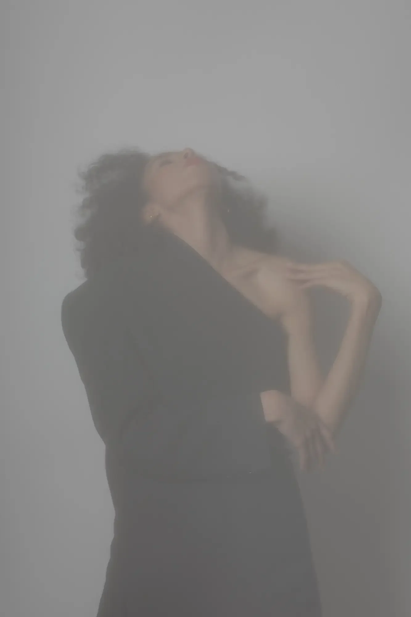 A woman with curly hair, dressed in a black outfit, standing behind a foggy or frosted glass, with her head tilted back and her hand resting on her shoulder.