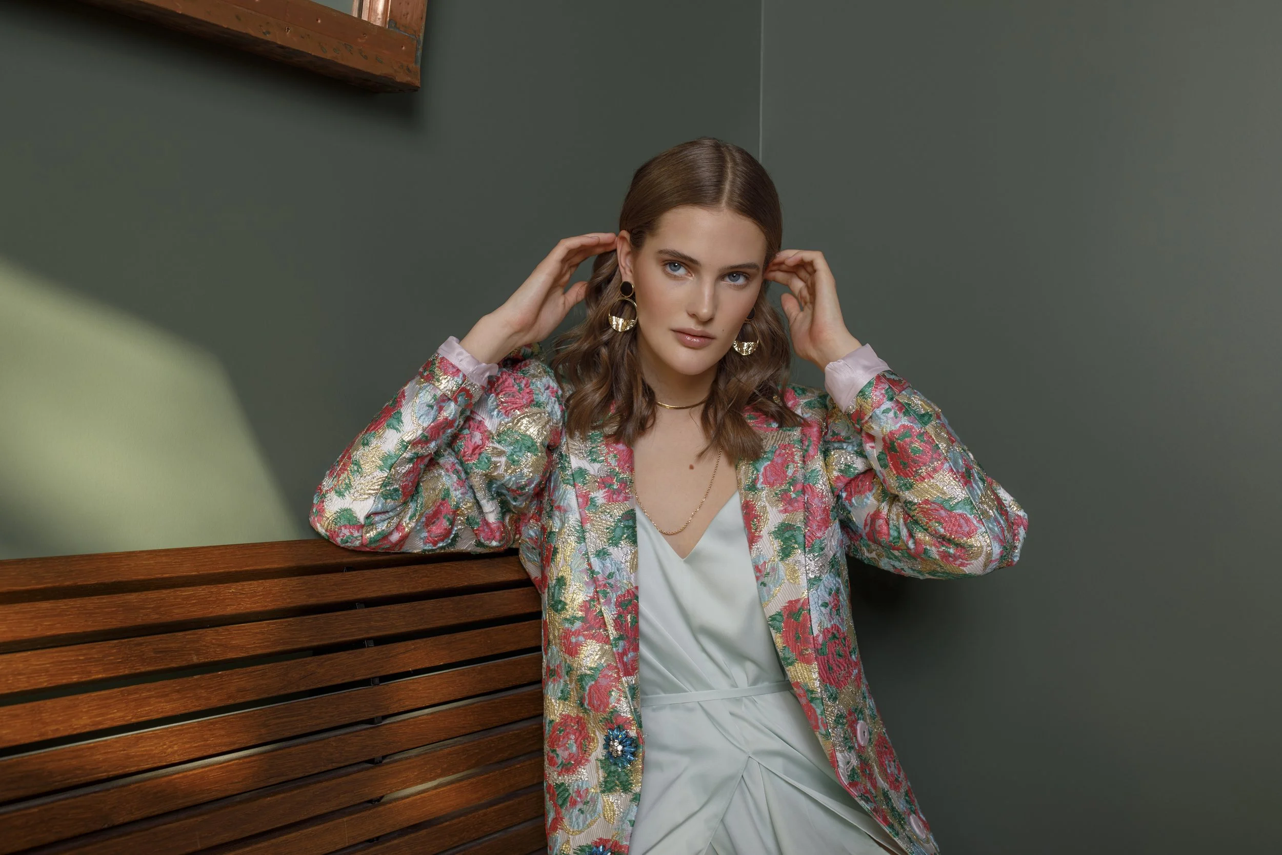 A woman with wavy brown hair wearing a floral patterned blazer and a white dress, sitting against a green wall with a wooden bench next to her, adjusting her earrings.
