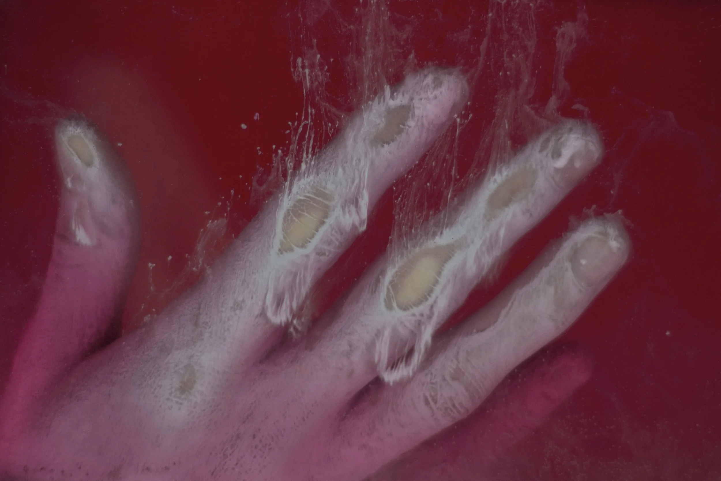 A hand submerged in a pinkish-red liquid, with soap bubbles and foam on the fingers and hand.