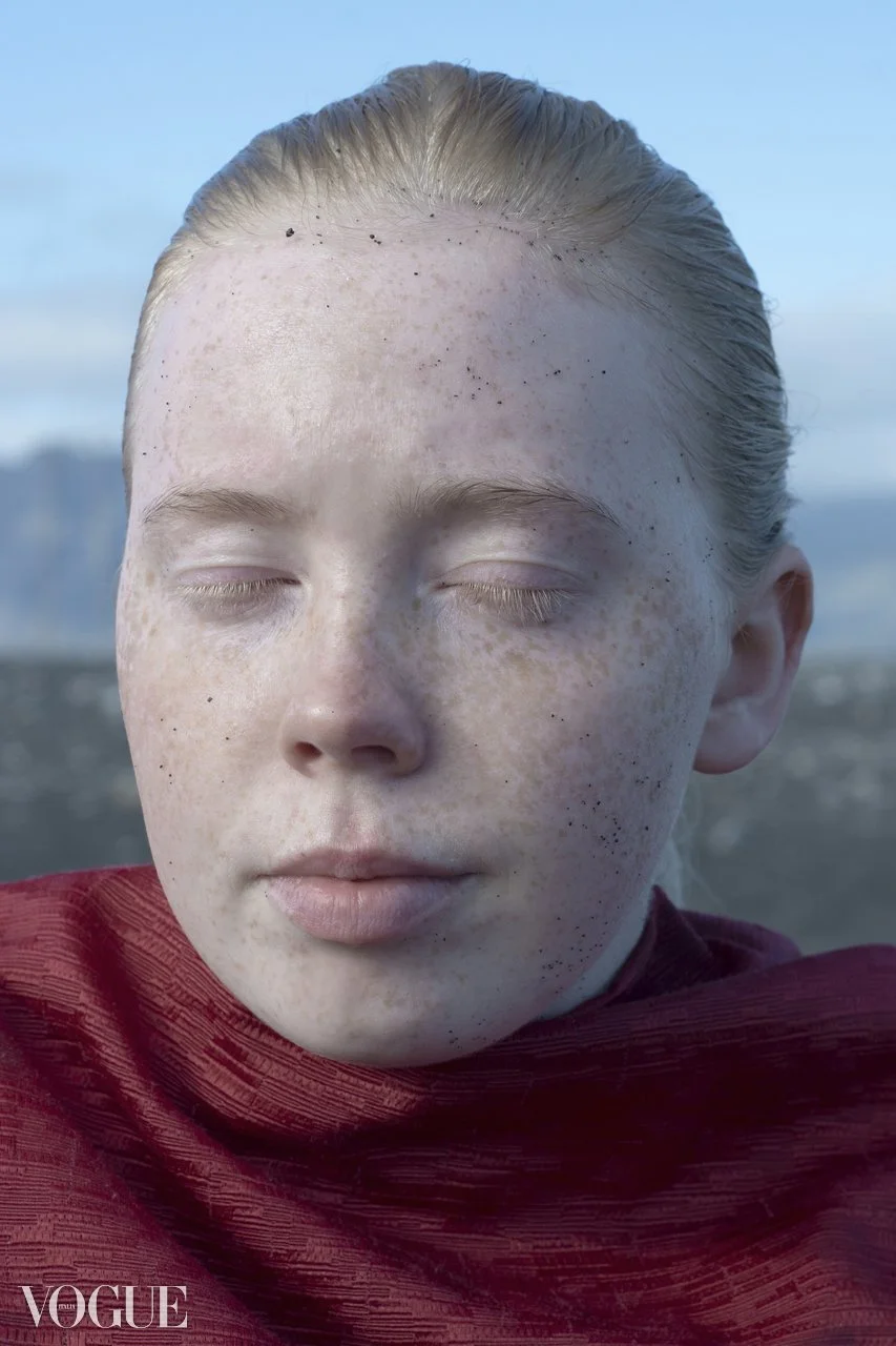 Close-up of a person's face with closed eyes, blond hair, and freckles, wearing a red scarf, outdoors with a blurred natural background.